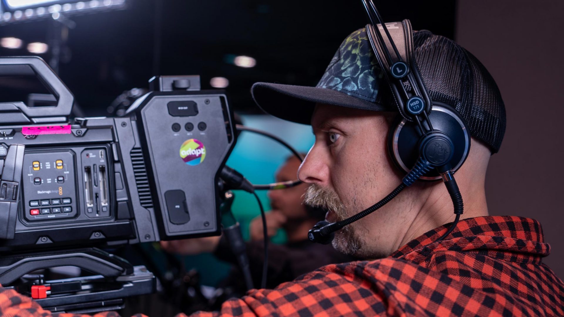A man wearing a headset and a cap operates a professional video camera in a studio, focusing intently. The camera has various controls and labels; studio lights and other equipment are visible in the background.