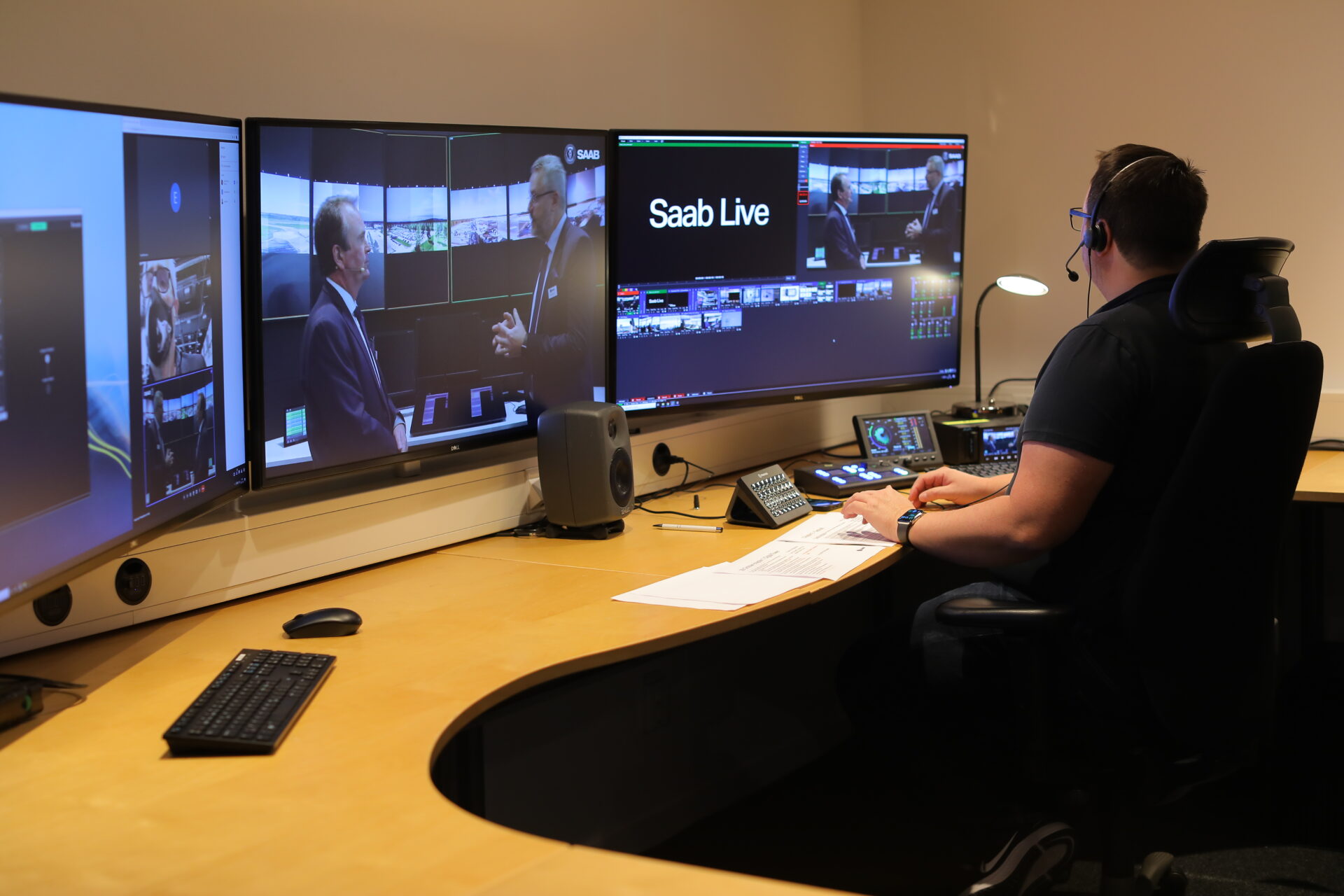 A person wearing a headset operates a multi-screen control station, monitoring a live broadcast with two people talking on the main screen labeled Saab Live in a modern studio environment.