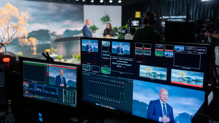 A live broadcast studio with monitors showing a man in a suit speaking against a scenic lake backdrop, while crew members manage the production equipment in the foreground.