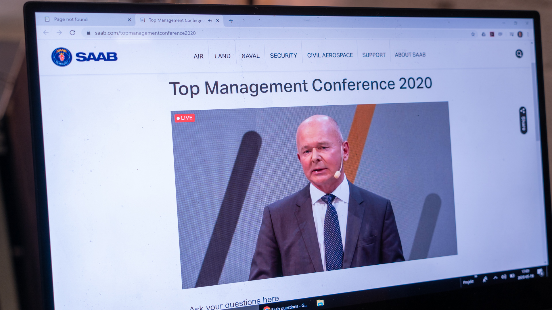 A computer screen displays the SAAB website showing a live stream of the Top Management Conference 2020, featuring a man in a suit speaking on stage.