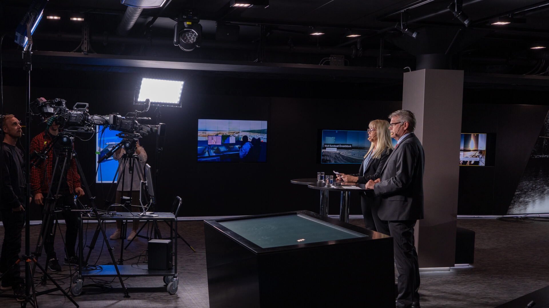 Two people in business attire stand at a podium, speaking on camera in a modern studio with professional lighting, monitors displaying graphics, and crew members operating equipment in the background.