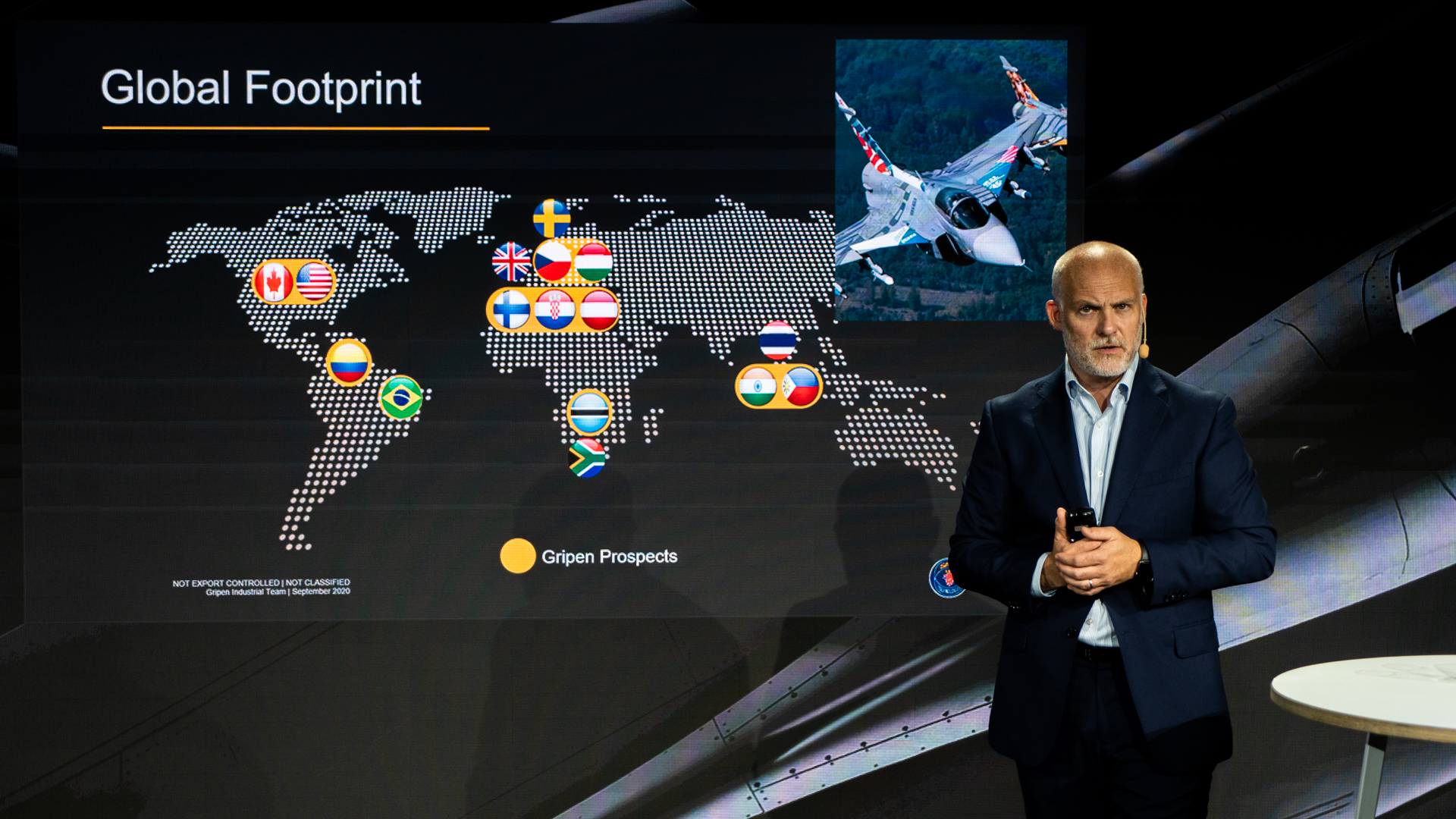 A man in a suit stands beside a screen showing a world map with various country flags marked as “Gripen Prospects,” and an image of a fighter jet in the top right corner. The title on the screen reads “Global Footprint.”.