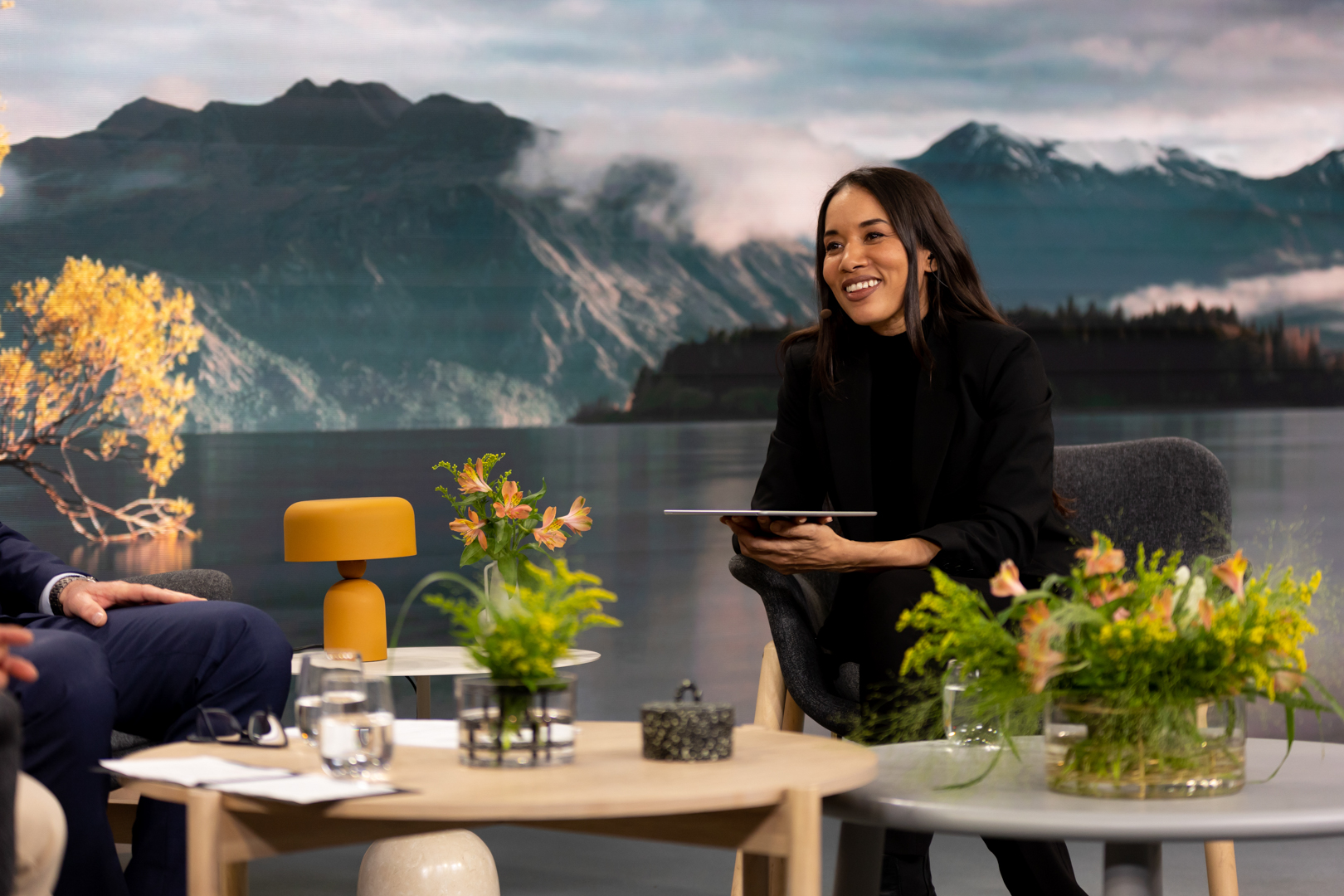 A woman in a black suit smiles while holding a tablet, seated in a modern studio with a mountain lake backdrop, surrounded by plants and a table with water glasses and decorative items.