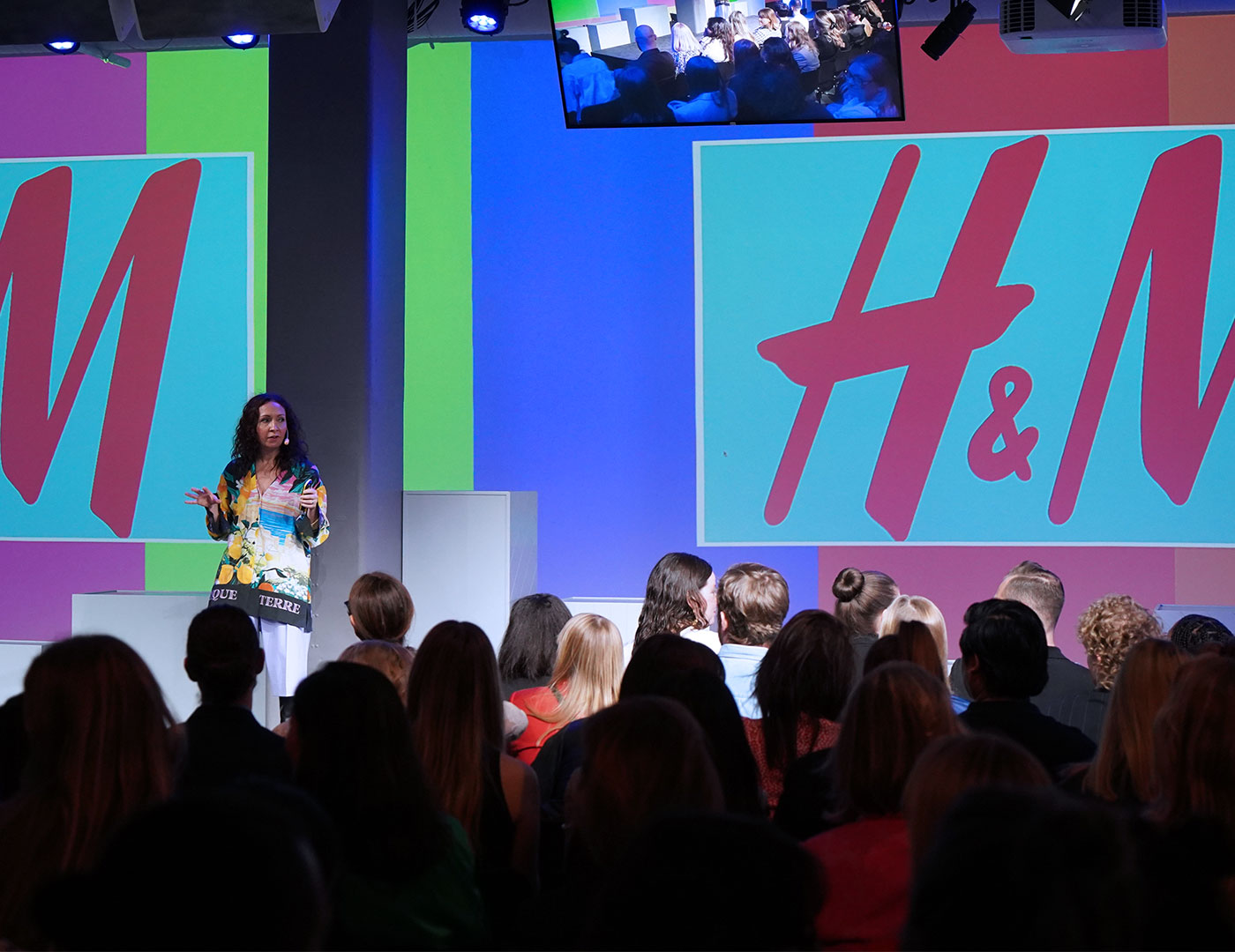 A woman speaks on stage to an audience at an event with large colorful H&M logos displayed on screens behind her and a small crowd image on a monitor above.