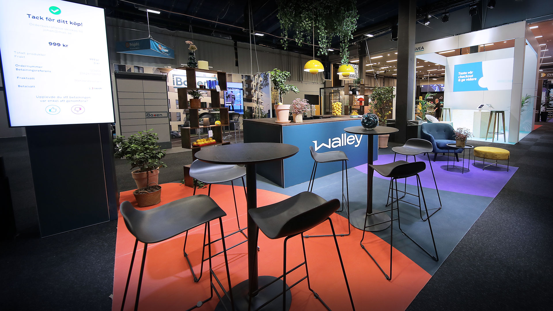 A modern exhibition booth with high tables and bar stools, plants, and colorful carpet sections. The booth features a counter labeled “Walley” and is set in a well-lit indoor event space.