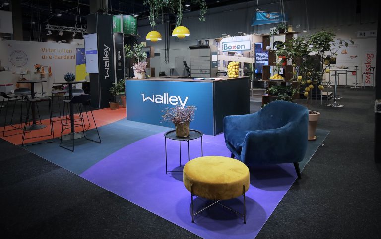A modern exhibition booth with Walley branding, featuring a blue chair, a yellow ottoman, a small table with a potted plant, and colorful flooring. Overhead, yellow lamps hang above a counter with lemons and greenery.
