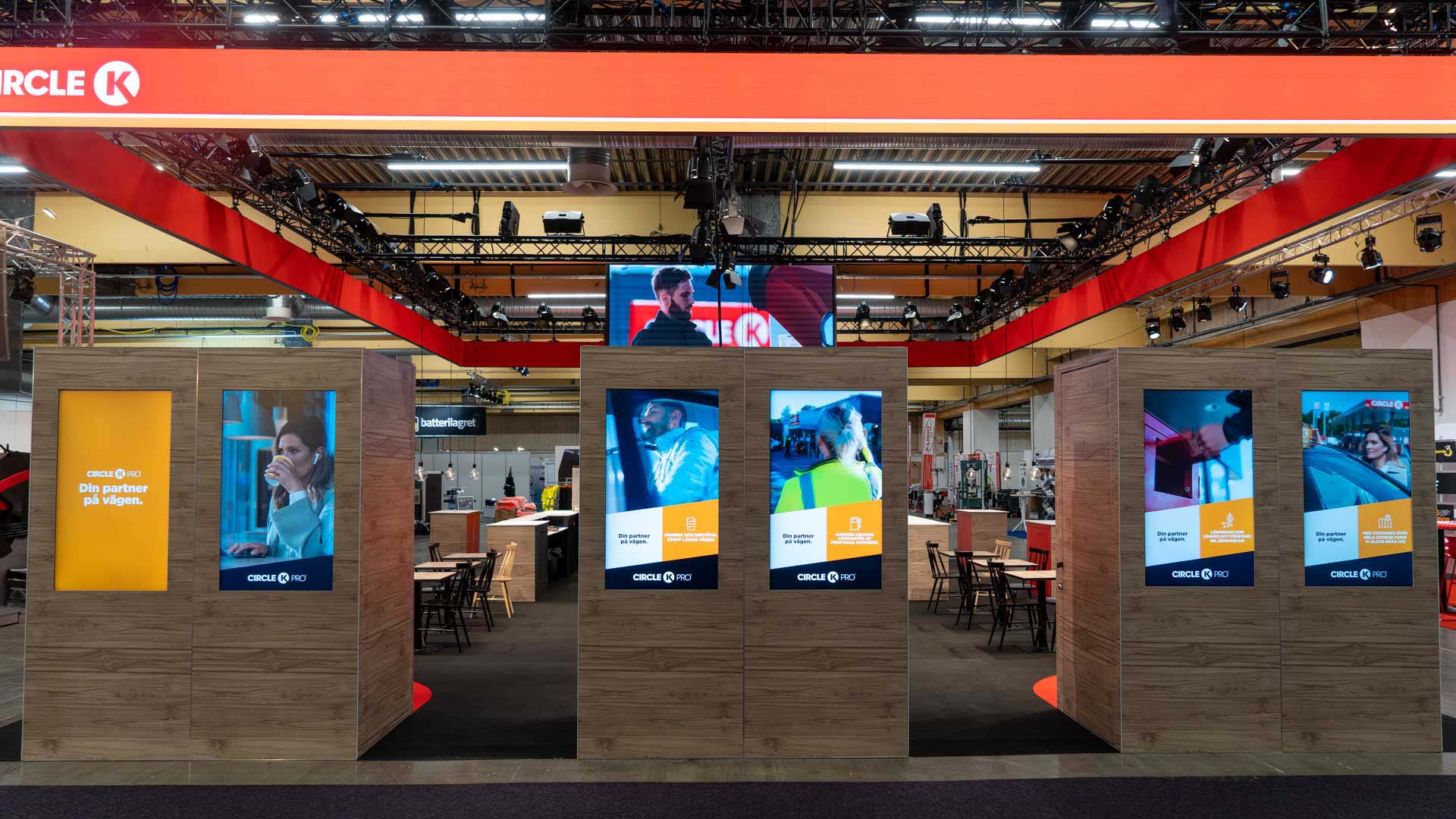 A trade show booth with wood-paneled displays featuring vertical digital screens showing various Circle K advertisements; tables and chairs are visible in the background under bright lighting.