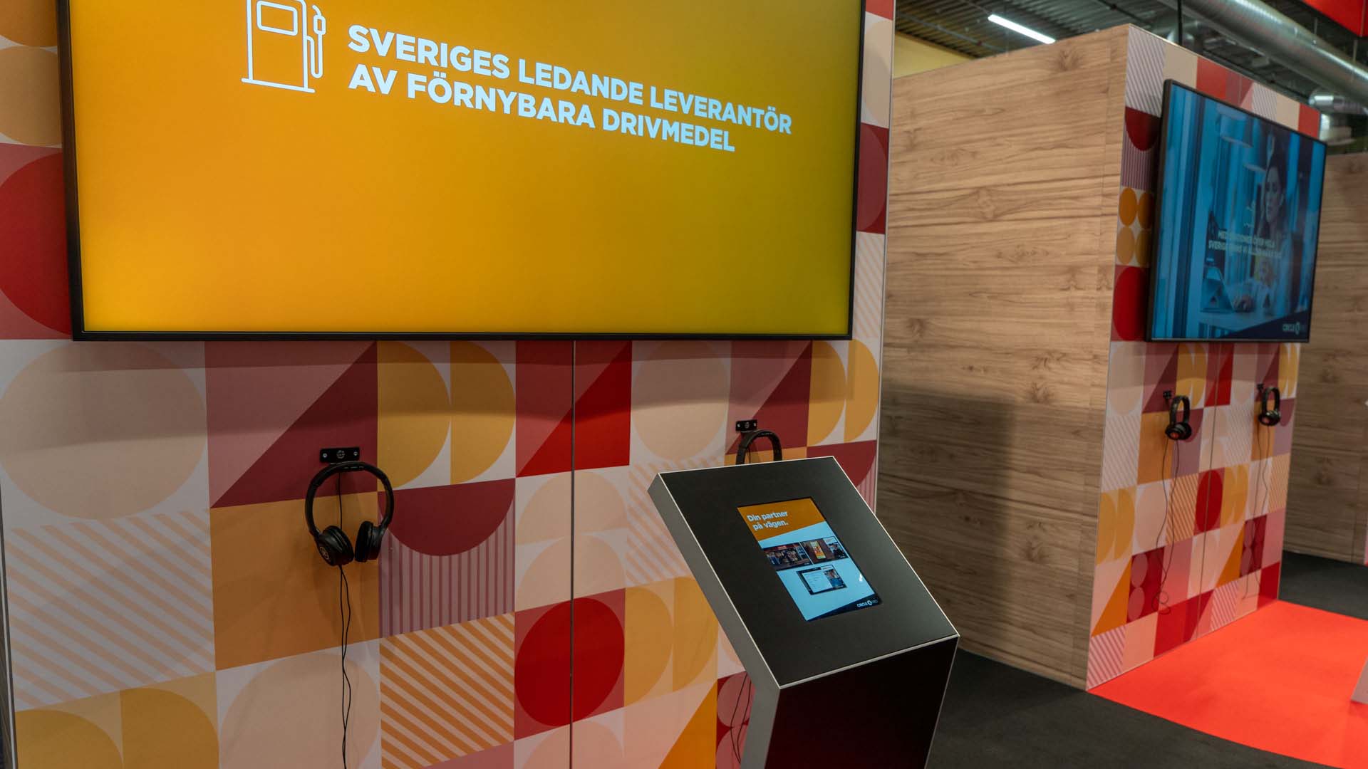 A digital kiosk with headphones is set up in front of a patterned wall and a large screen displaying Swedish text about renewable fuels at an indoor exhibition booth.