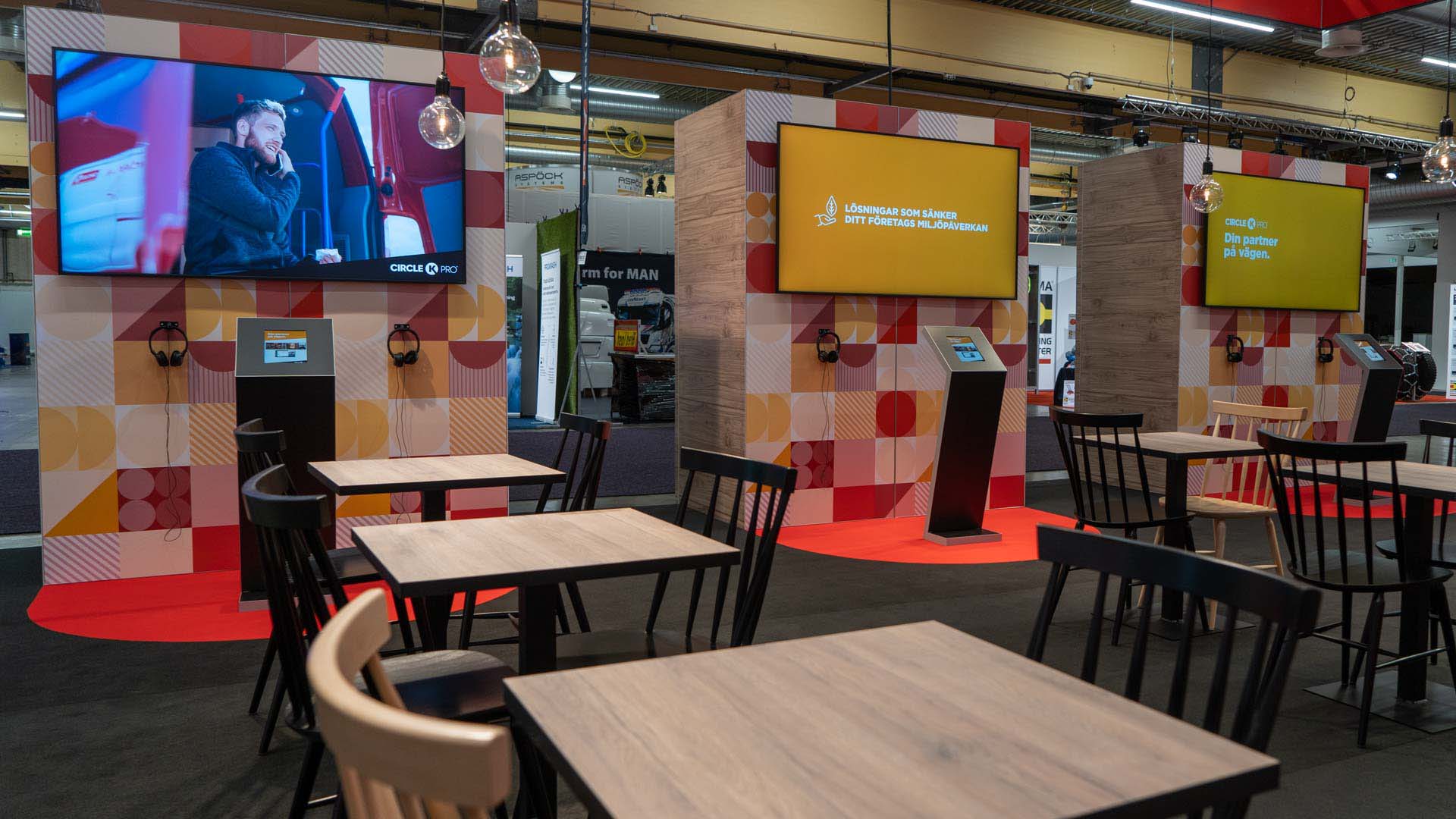 A modern exhibition booth features tables and chairs in the foreground, two large screens with colorful graphics and text, and a TV screen displaying a person talking. Two kiosks stand between the screens.