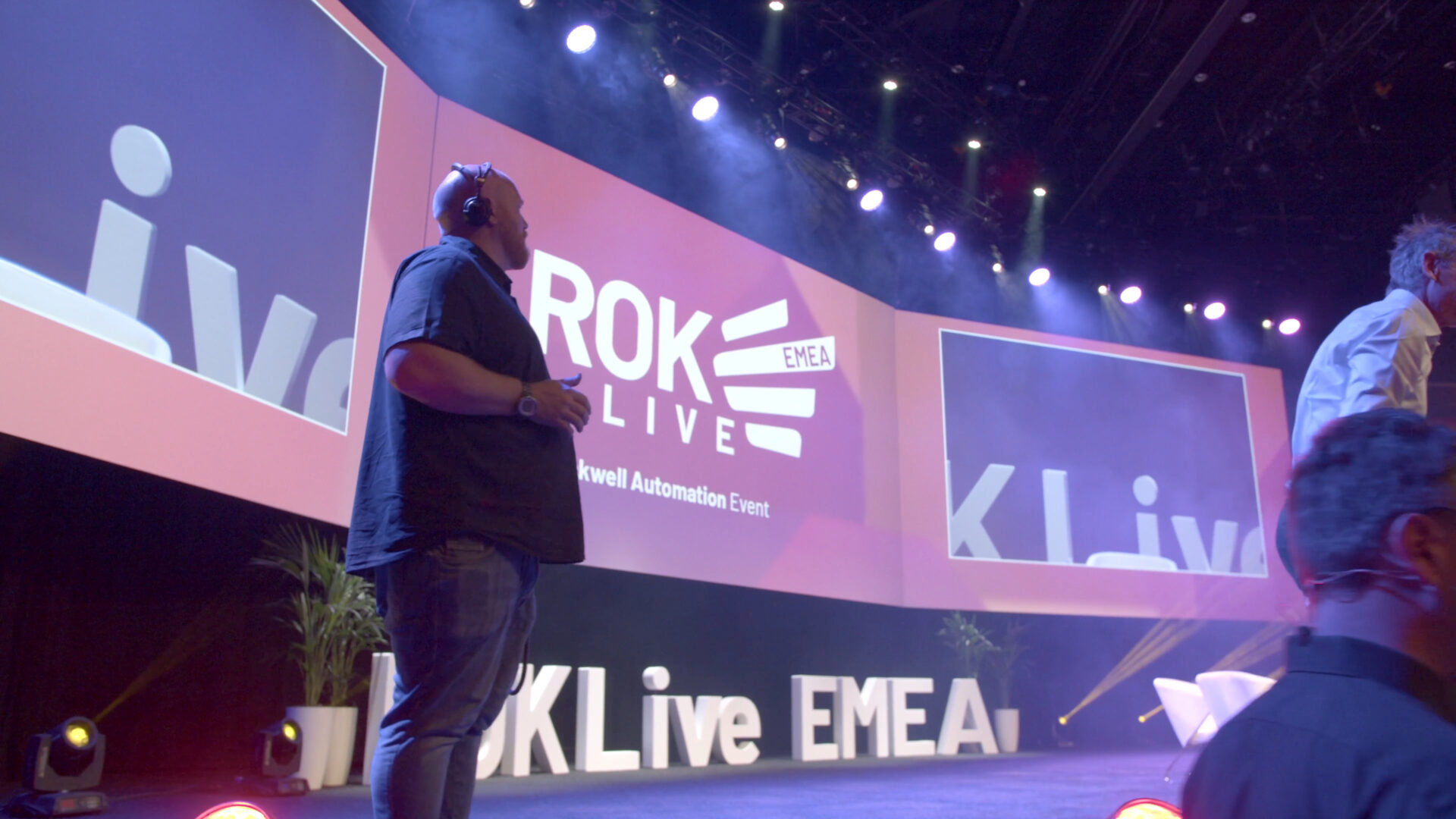 A person stands on stage at the ROKLive EMEA Rockwell Automation Event, with large screens and stage lights in the background displaying the event branding.