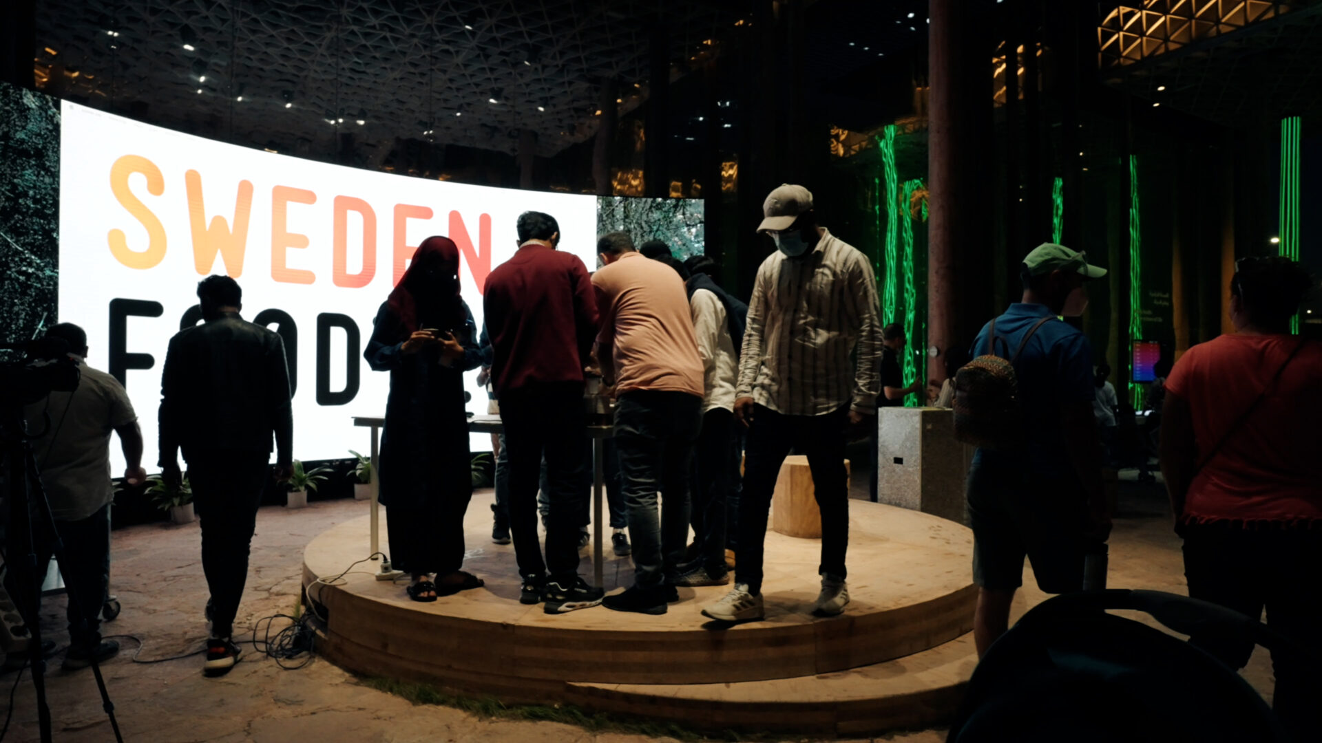 A group of people stand and walk on a small round stage in front of a large illuminated sign that reads SWEDEN FOOD. The background shows a modern, indoor space with green lighting.