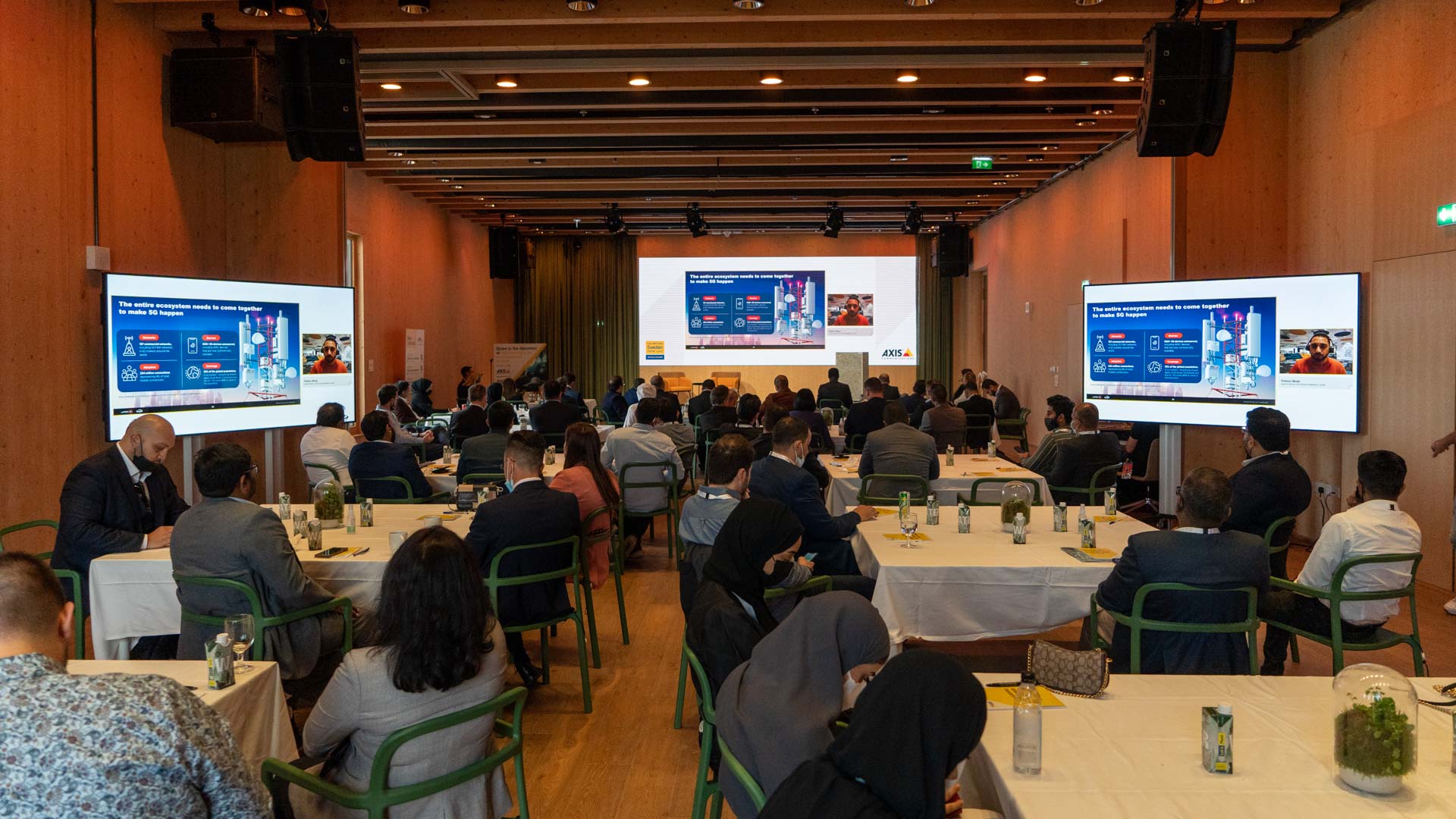 Conference attendees sit at tables in a modern hall, facing large screens displaying presentation slides with graphics and text. The room has wooden walls, green chairs, and bottles of water on each table.