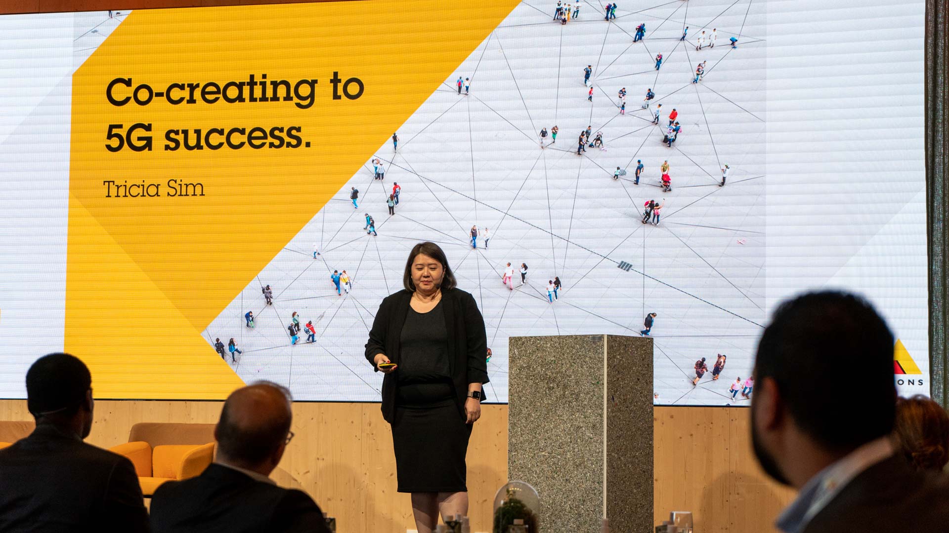 A woman stands on stage in front of a large screen displaying the text “Co-creating to 5G success. Tricia Sim” alongside an aerial image of people walking. Audience members watch from their seats.