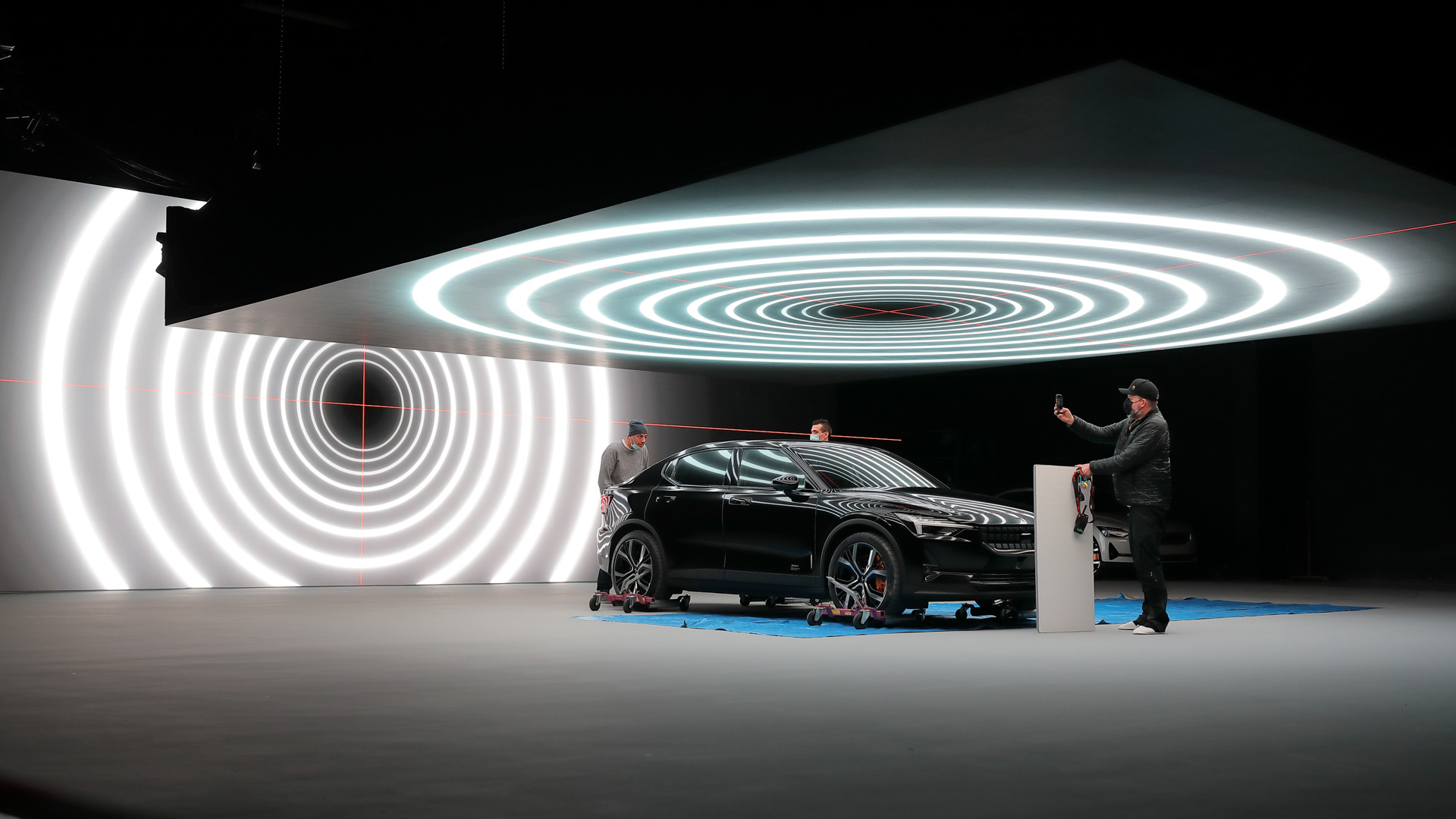 A sleek black car is displayed indoors under bright, concentric circular lights on the ceiling and wall. Three people surround the car; one person appears to be taking a photo with a smartphone.