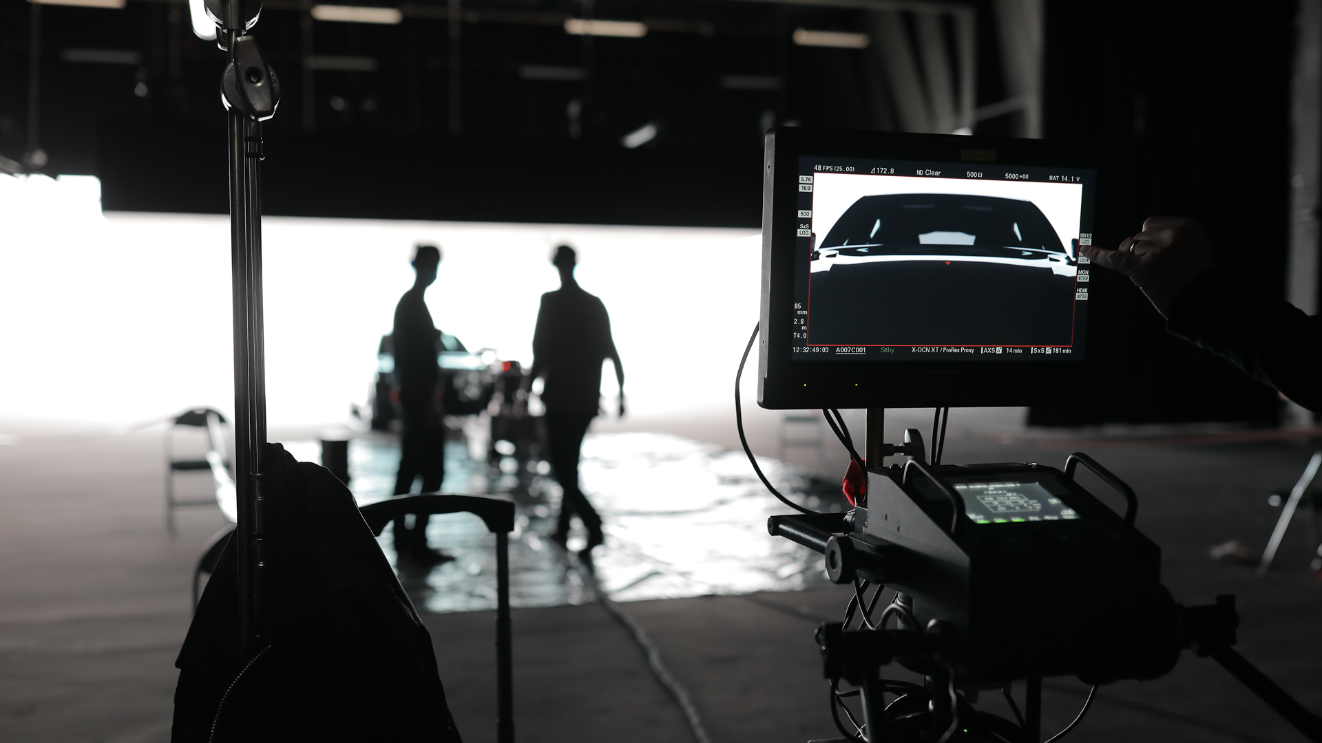 A film set with a monitor displaying a car scene, while two people stand blurred in the background near a vehicle, illuminated by bright backlighting in a dark studio.