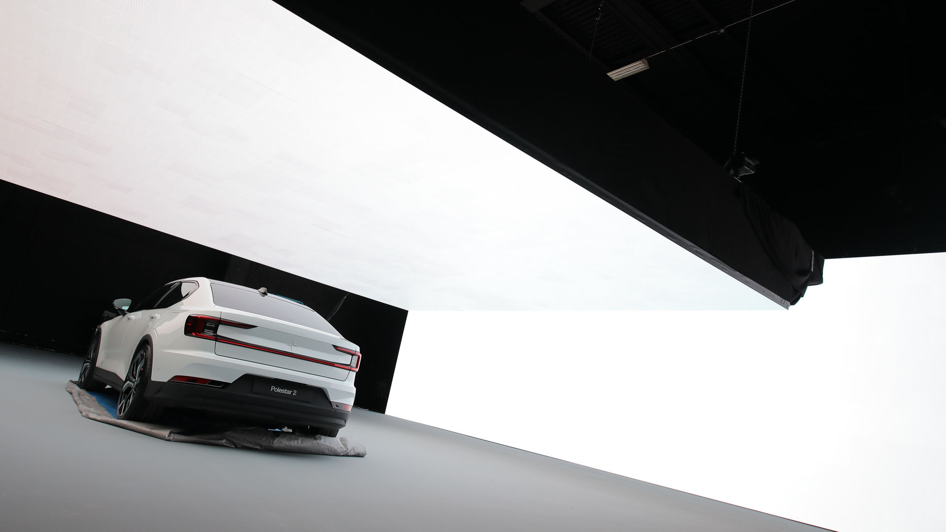 A white car is parked indoors on a large gray floor, partially covered by a cloth. The background is minimalistic with bright white and black contrasting walls and ceiling panels.