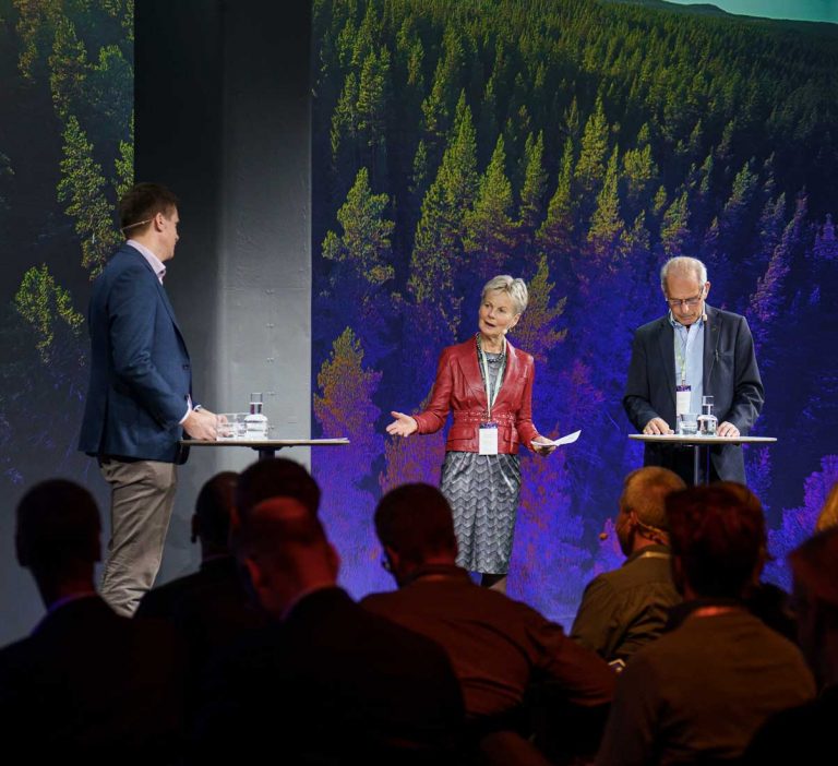 Three people are standing at tall tables on a stage, engaged in discussion, with an audience in front of them. The background features a large, vibrant image of a forest.