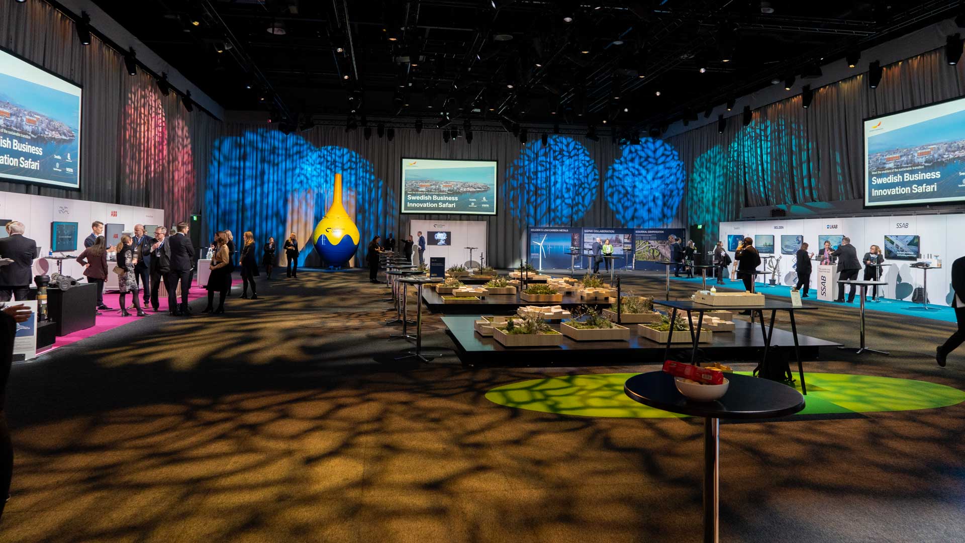 A spacious event hall with people networking near exhibition booths, buffet tables with food, and colorful, leaf-patterned lighting projected on walls and floors. Large screens display “Swedish Business Innovation Safari.”.