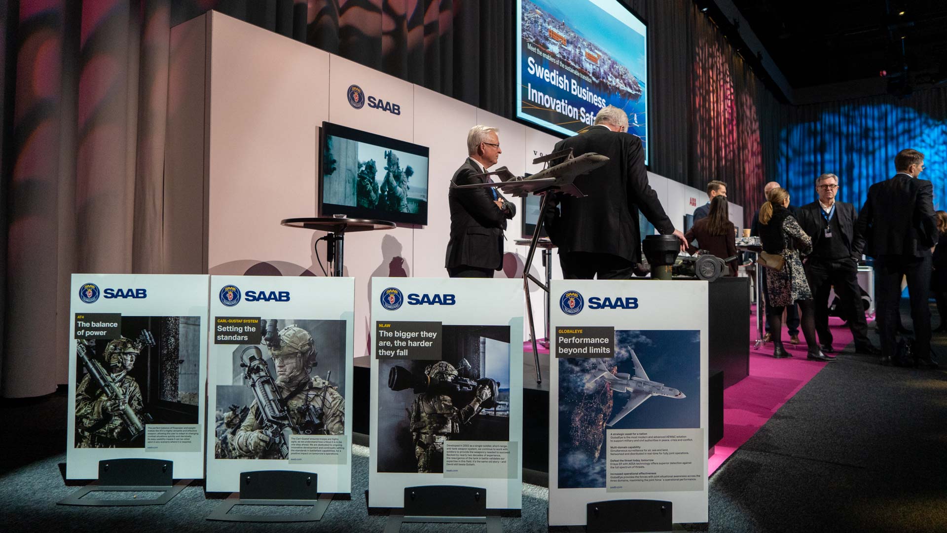 A group of people in business attire interact at a Saab exhibition booth with military-themed displays, posters, and models, under bright lighting at a convention or conference.