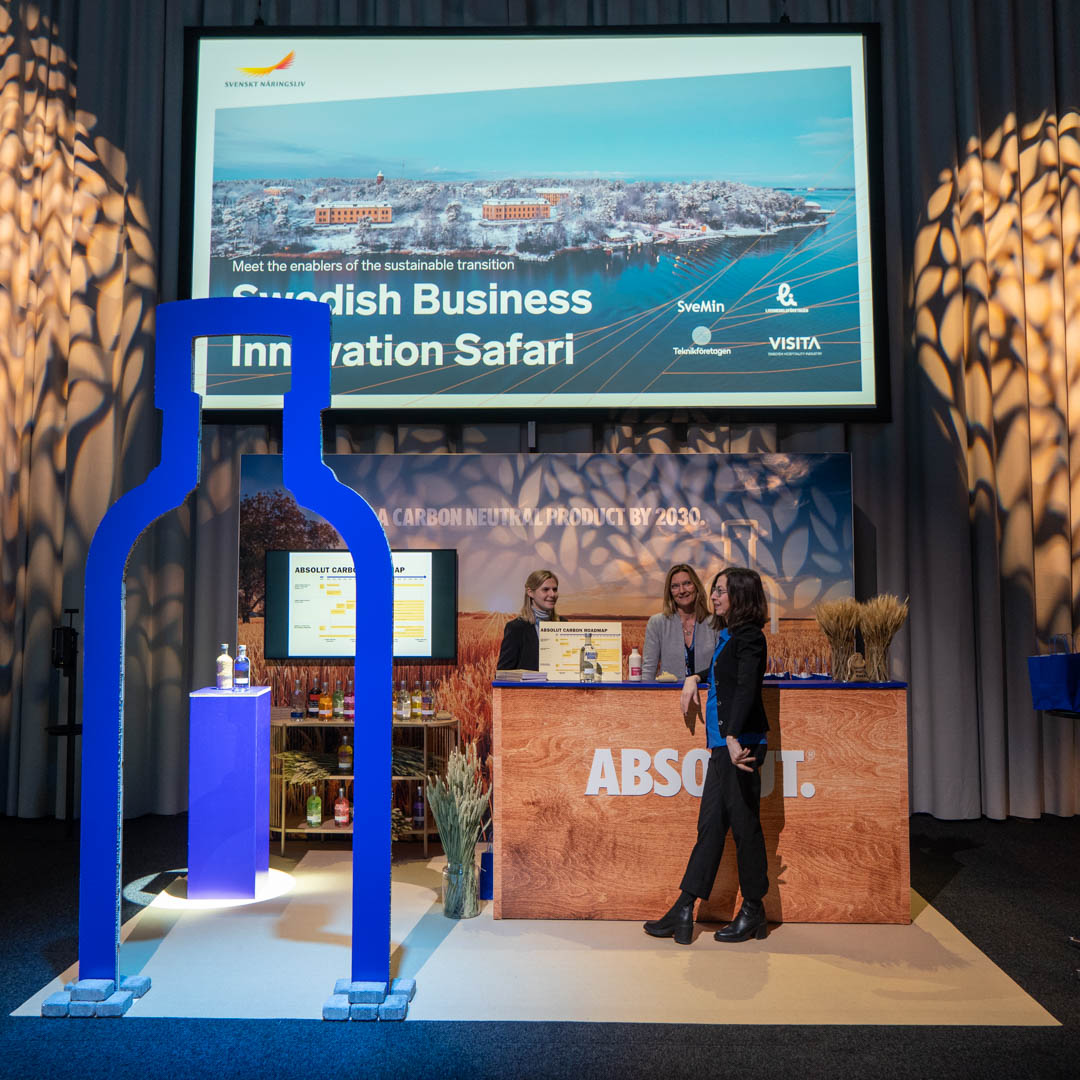 Exhibit booth with a large blue bottle-shaped frame, three people interacting at a counter with “ABSOLUT” branding, and a screen displaying “Swedish Business Innovation Safari.” Wheat decorates the booth.