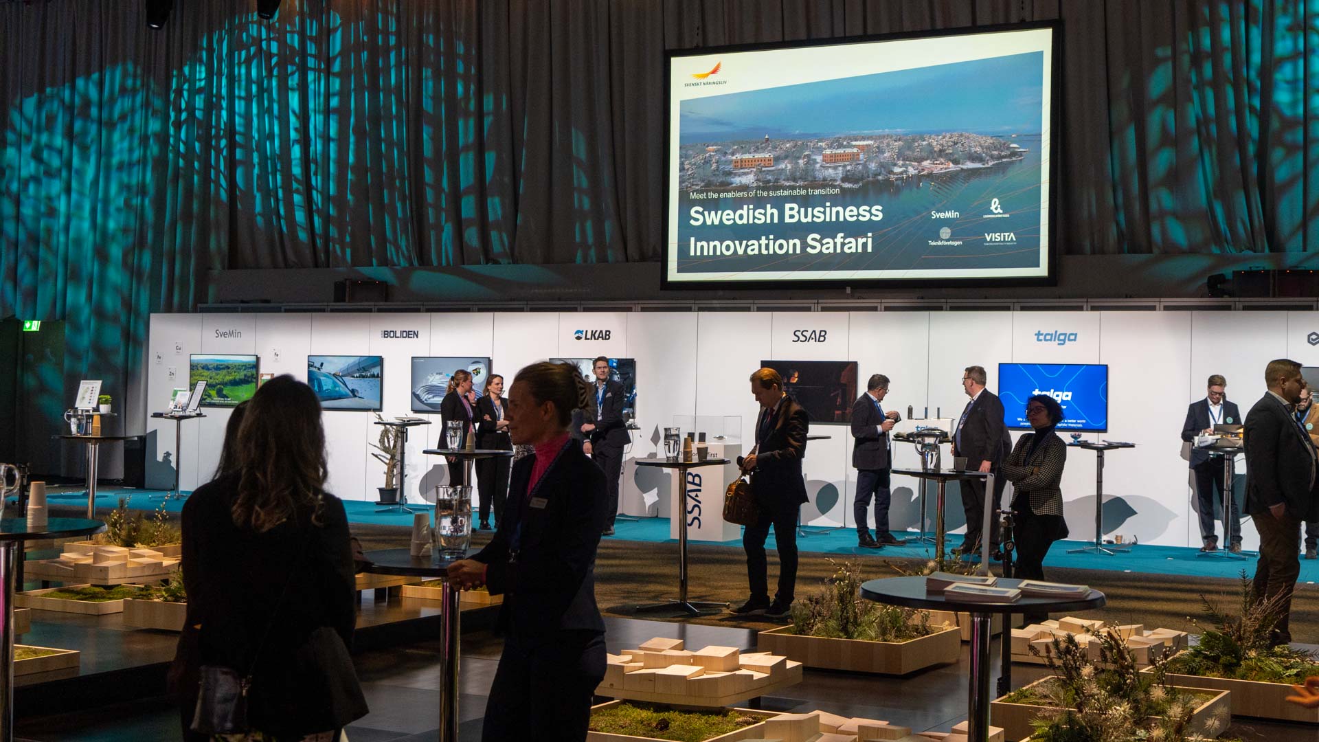 People networking at an indoor business event with booths. A large screen displays “Swedish Business Innovation Safari” above branded stands. The venue is modern, with blue lighting and minimalist decor.