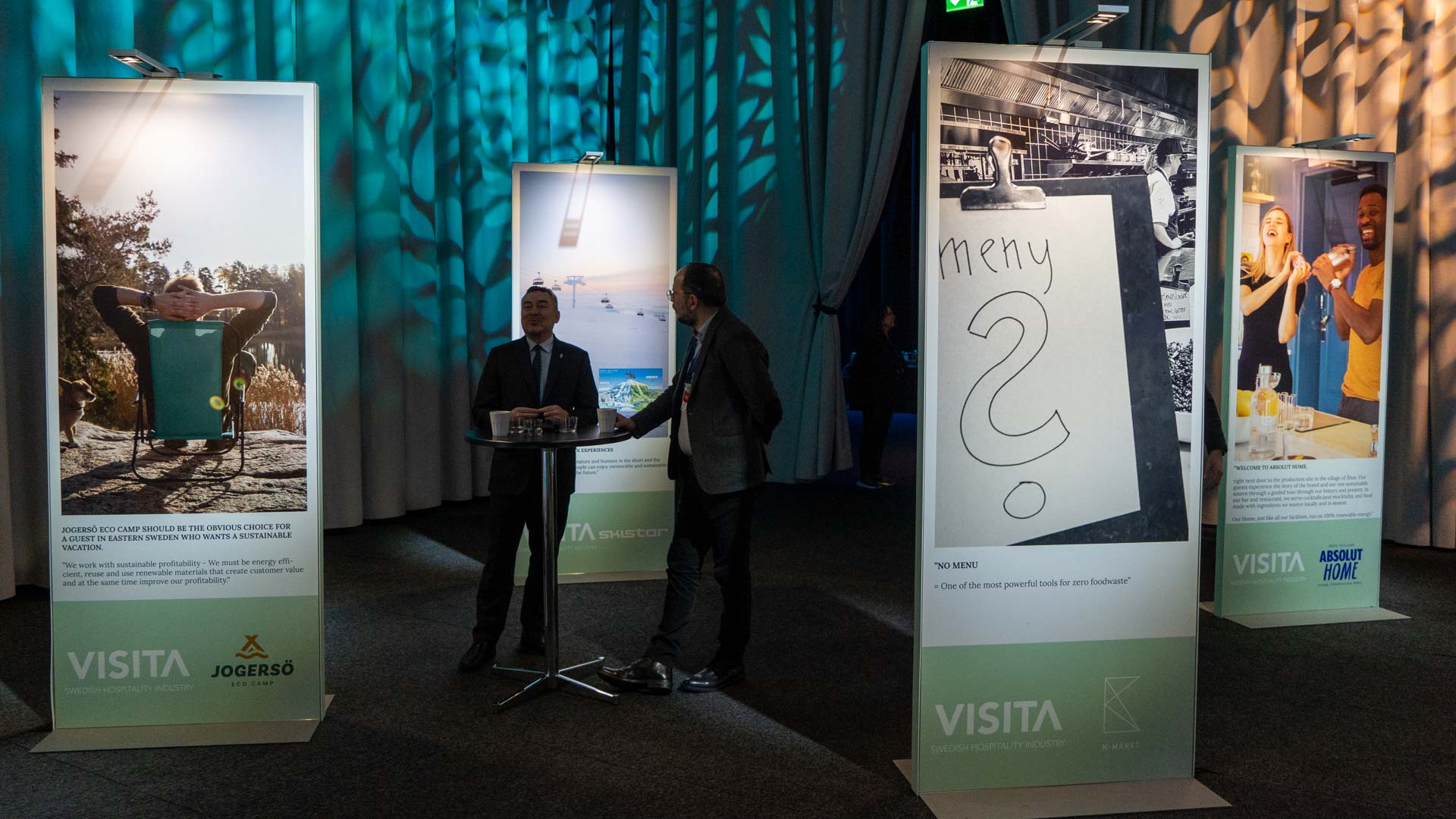 Two men stand and talk at a small round table in a dimly lit room with large, illuminated display panels showing photos, drawings, and text. The environment appears to be an exhibition or event.