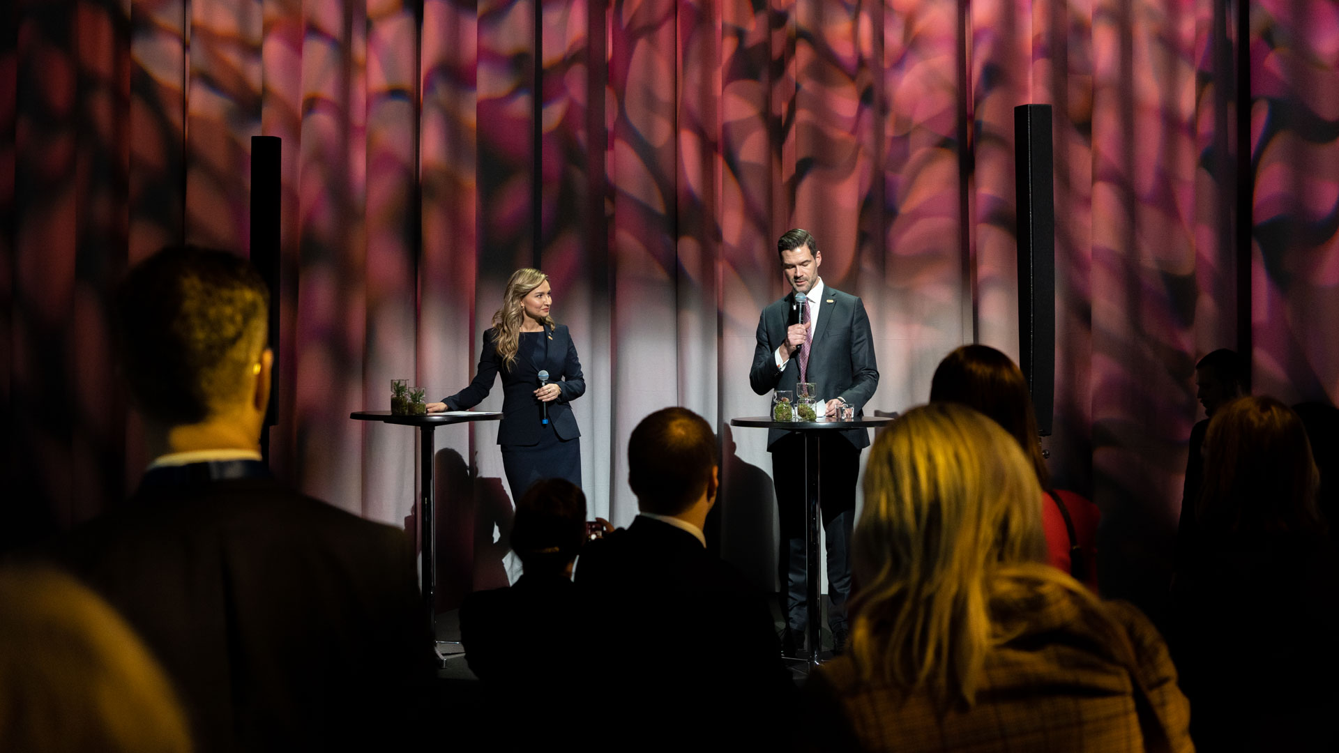 Two speakers, a woman and a man in formal attire, stand at separate podiums on stage addressing an audience. The background features dramatic, patterned lighting with shades of pink and purple.
