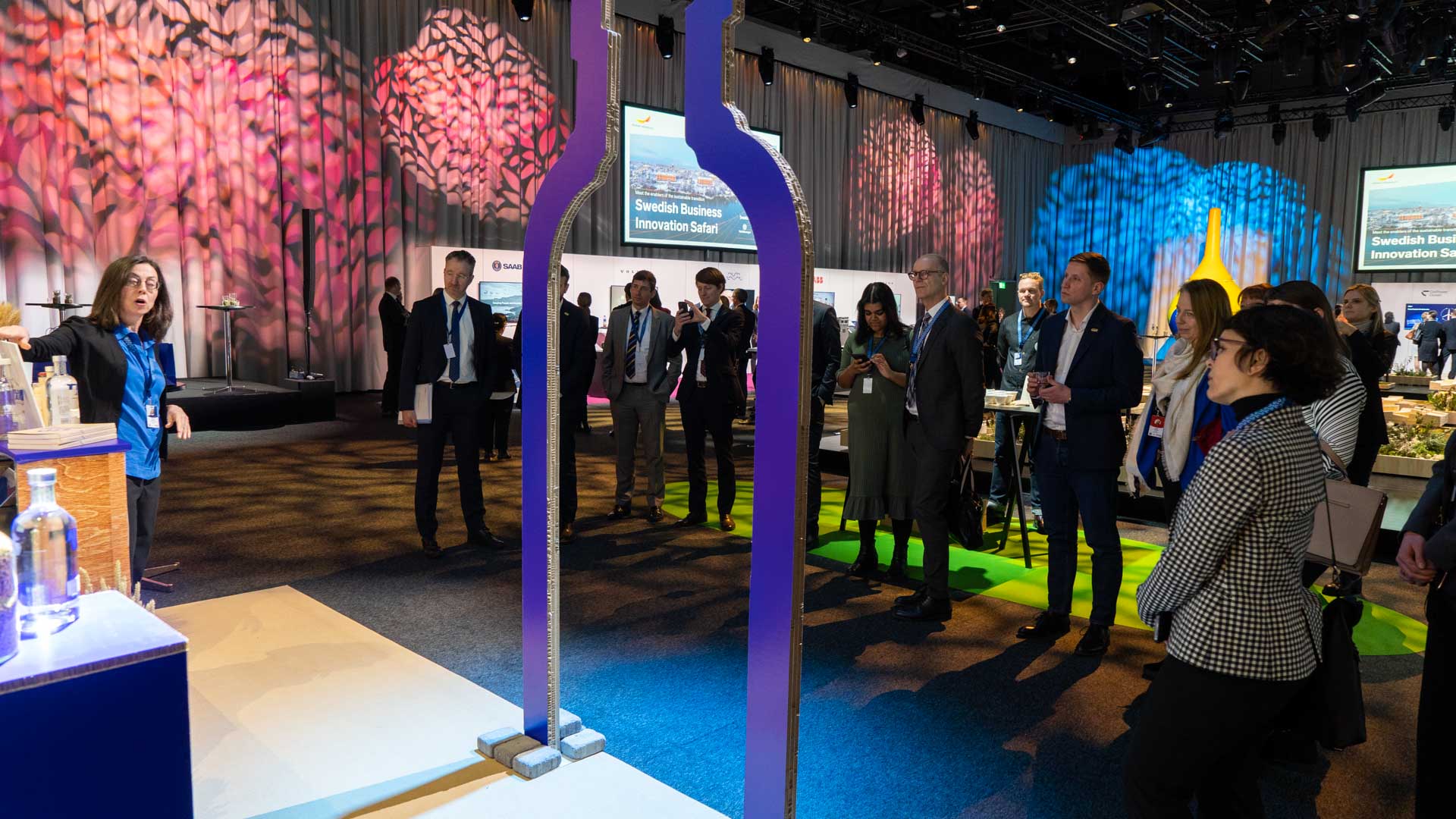 A woman presents to a group of people at an indoor event, with decorative lighting on curtains and displays on tables. The audience listens attentively, and two large purple frames stand in the foreground.