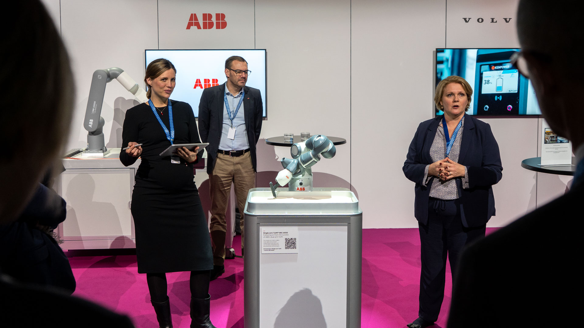 Three people stand in a booth with ABB branding, presenting a small robotic arm on a pedestal. Two display screens and a gray robotic arm are visible in the background, and the floor is bright pink.