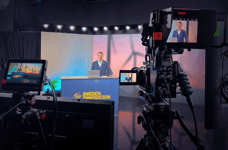 A man sits at a desk with a laptop, hosting a show called Sweden Innovation in a studio with cameras, lights, and monitors displaying his image from different angles.