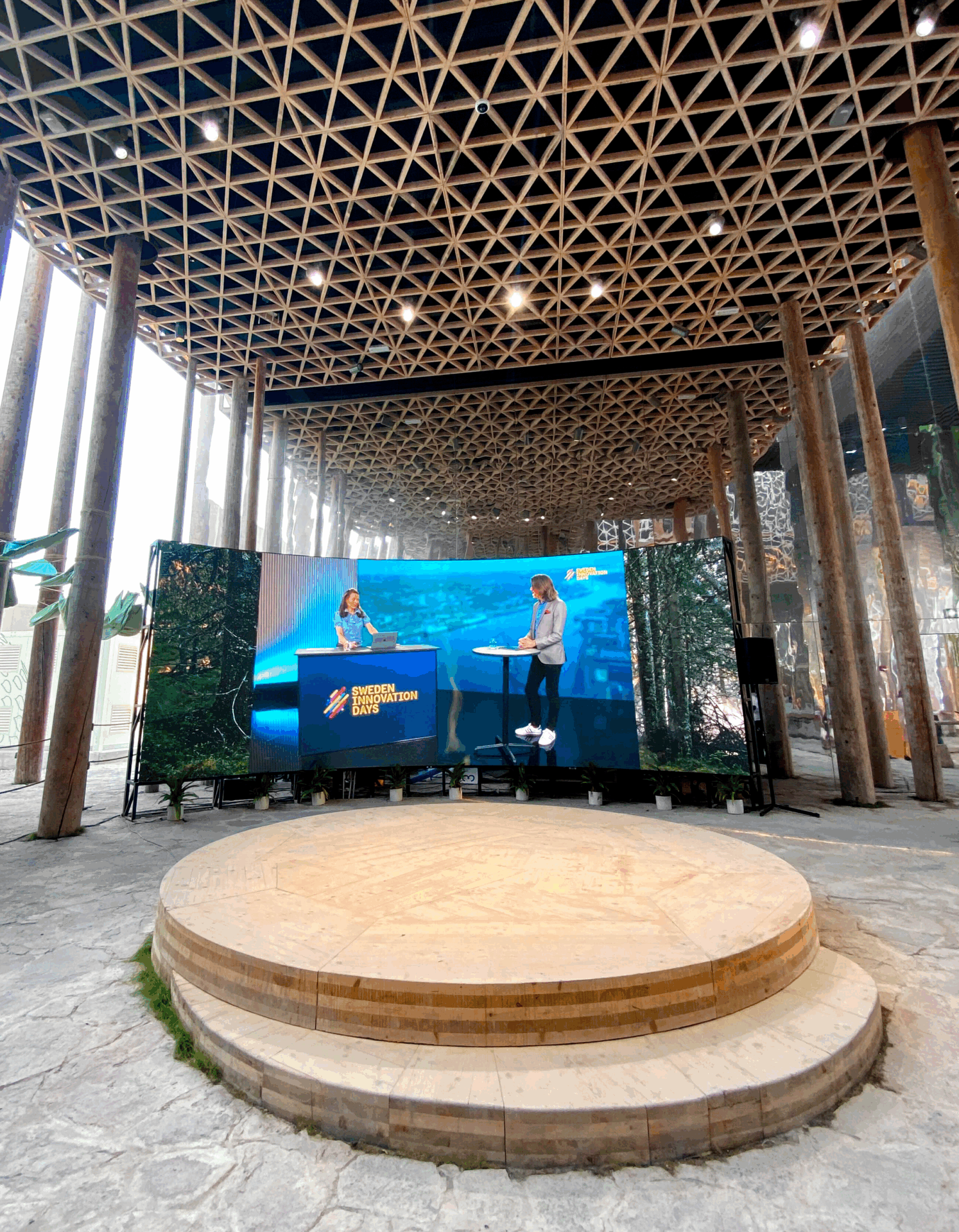 A circular wooden stage sits in front of a large screen displaying two people speaking at an event called British Innovation. The venue has tall wooden columns and a geometric ceiling.