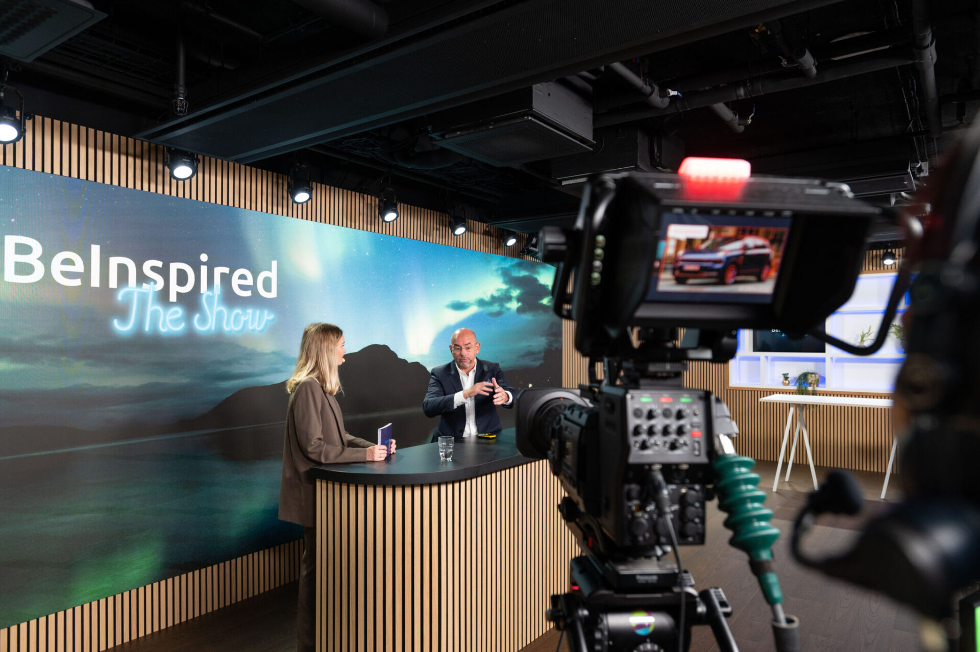 A camera films two people having a conversation at a modern studio desk with a “BeInspired The Show” sign and scenic backdrop, while studio lights and equipment are visible in the foreground.