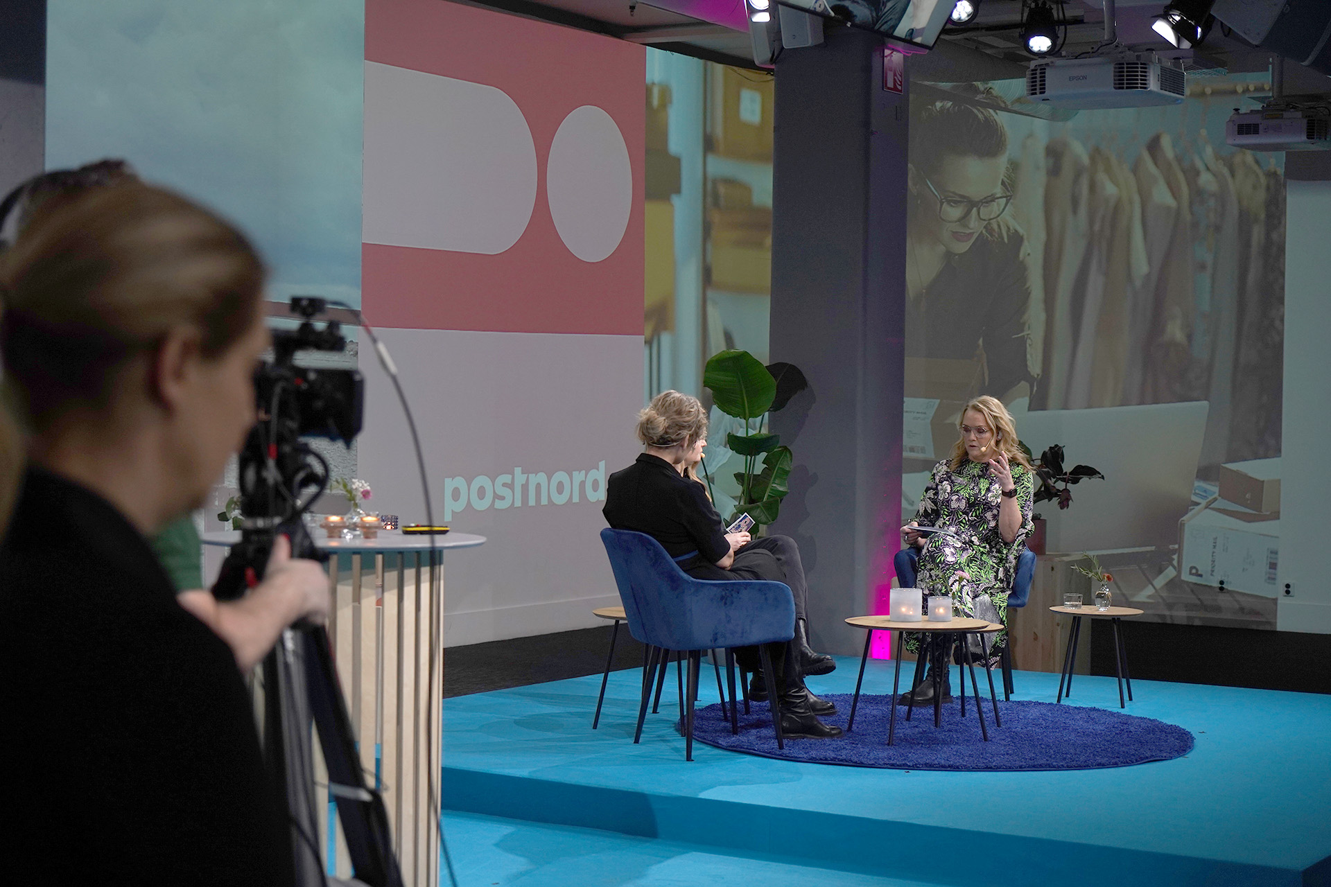 Two women sit on a small stage having a discussion, with one person filming them. Behind them is a large screen displaying the PostNord logo and an image of a woman working on a laptop in a clothing store.