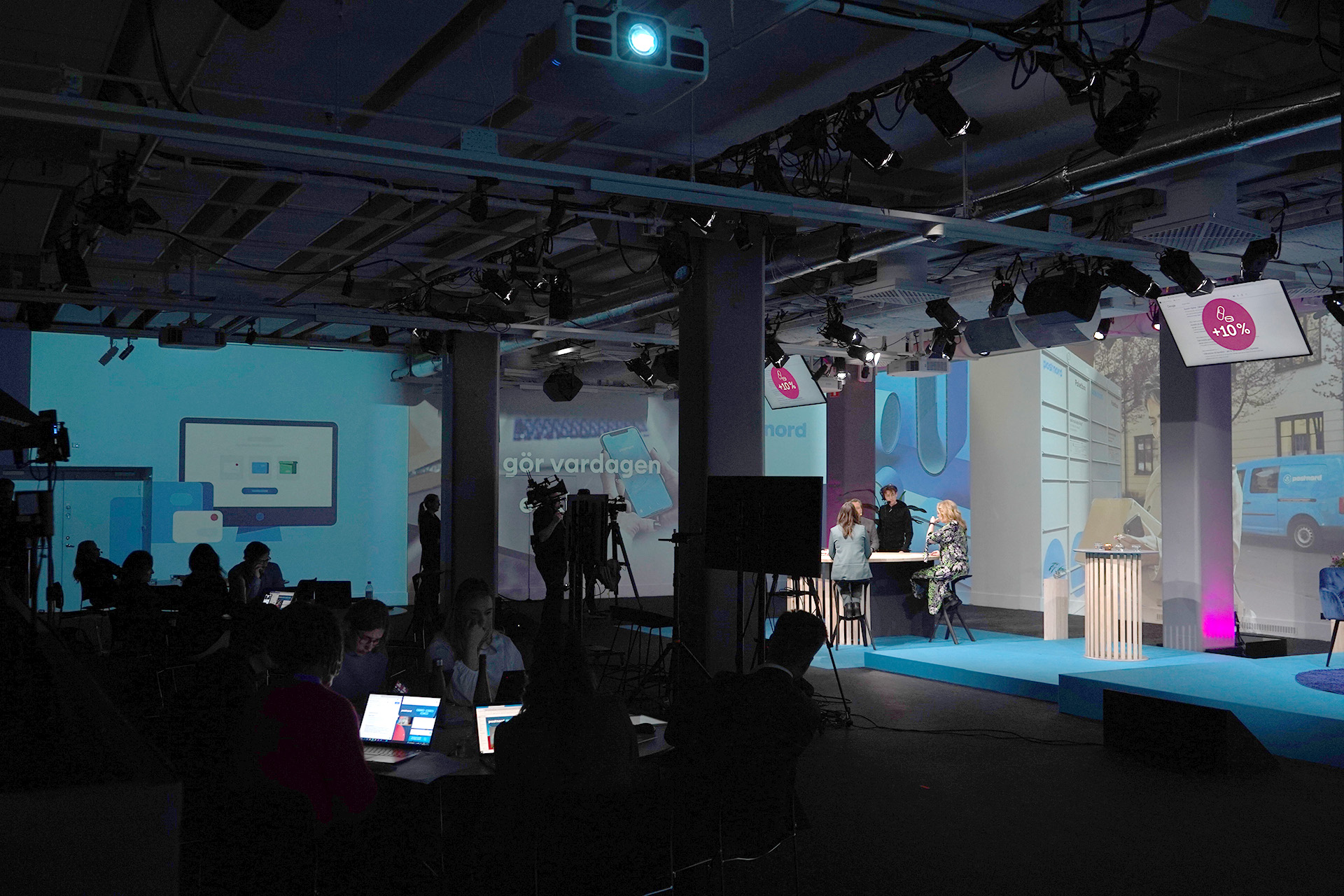 A modern conference room with people working on laptops in dim lighting while a panel of speakers talks on a brightly lit stage with screens displaying graphics and text in Swedish.