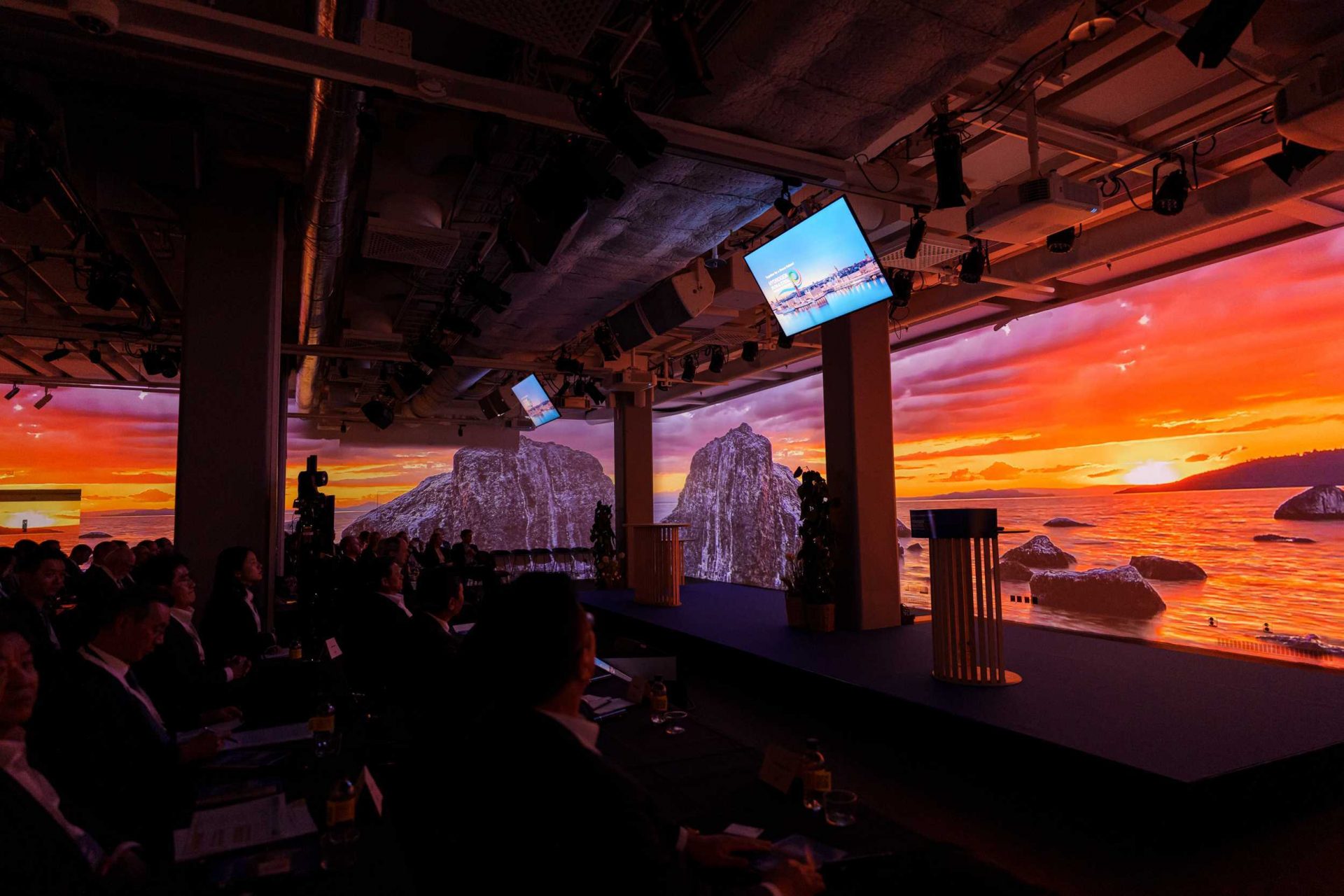 A conference room with people seated in the audience, facing a stage and large digital screens displaying a vivid sunset over rocky seashores. Monitors hang from the ceiling, and the atmosphere is modern and immersive.