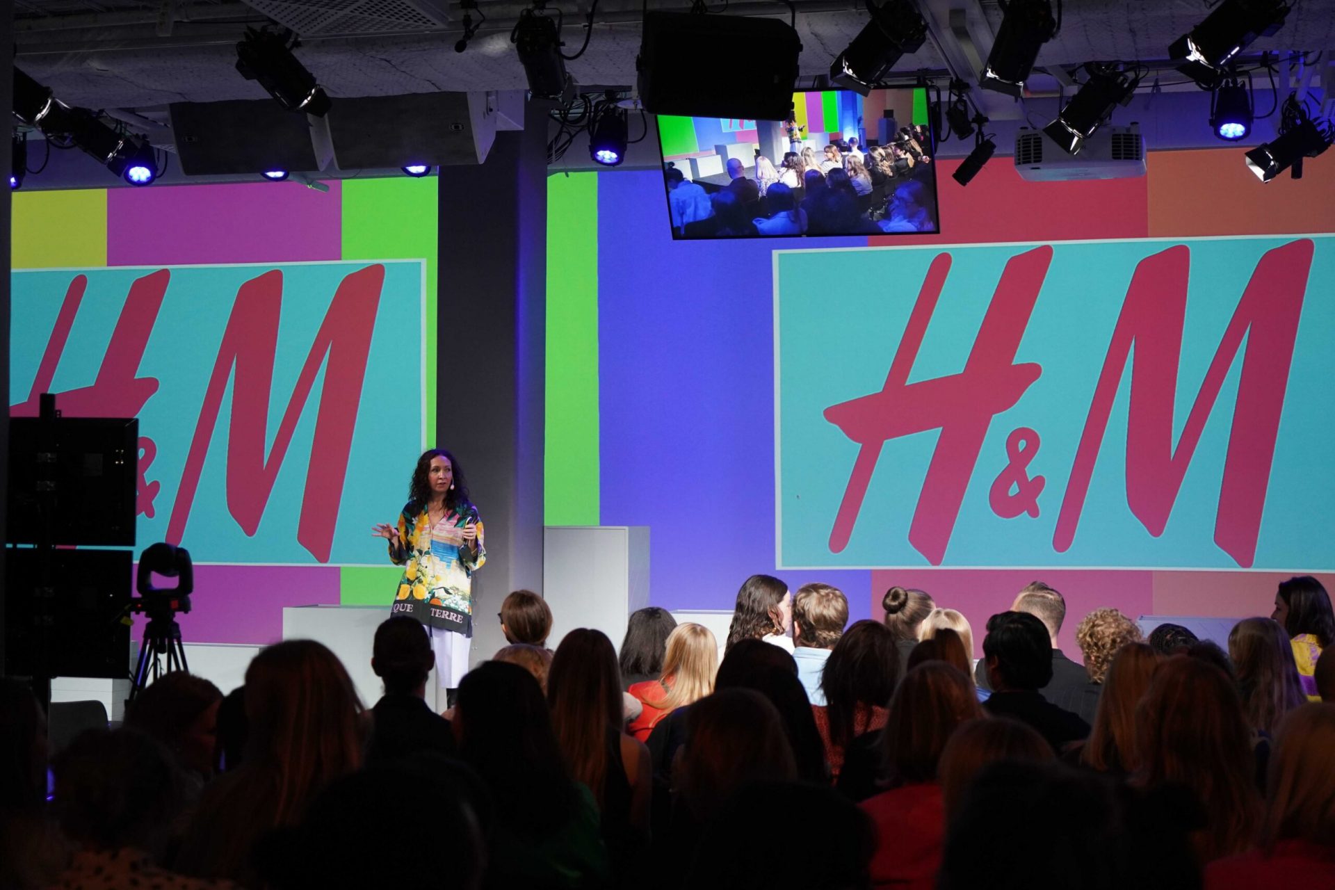 A woman speaks on stage at an H&M event in front of a seated audience, with large colorful H&M logos displayed on screens behind her.