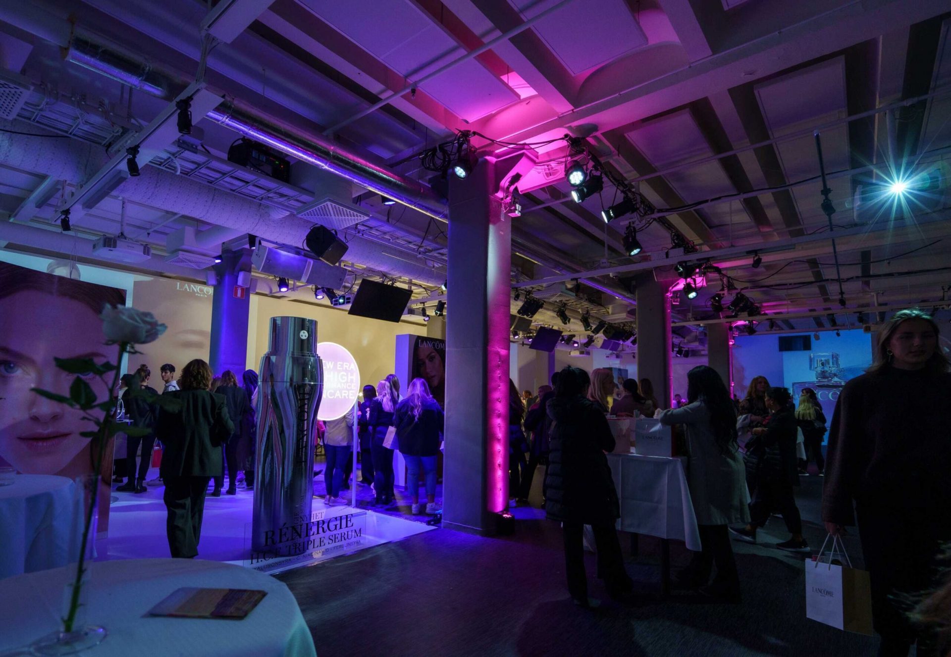 A crowd of people socialize at an indoor event with purple and blue lighting, surrounded by branded displays and large images of a woman’s face. Cocktail tables and event decorations are visible throughout the space.