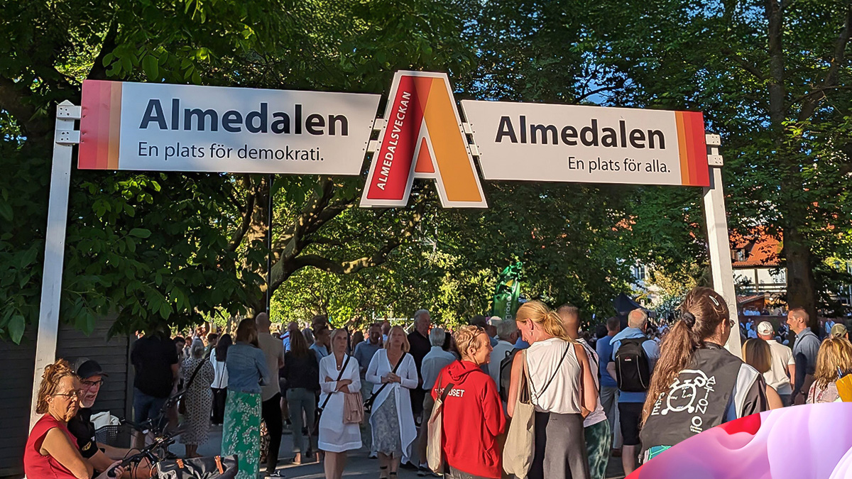 A crowd gathers under signs reading Almedalen – A place for democracy and Almedalen – A place for everyone at the entrance to an outdoor event, surrounded by trees and summer sunlight.