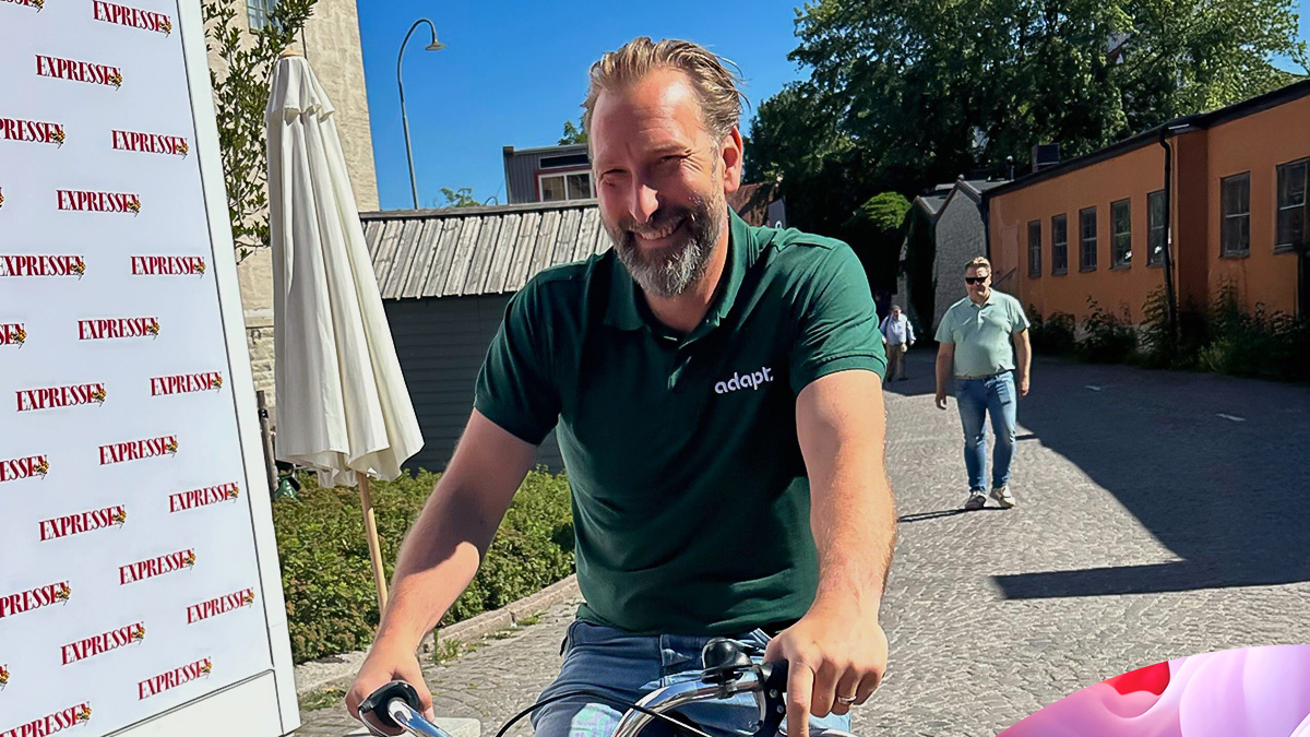 A smiling man with a beard, wearing a dark green polo shirt, rides a bicycle on a sunny cobblestone street. Another man walks behind him. Buildings and trees are visible in the background.