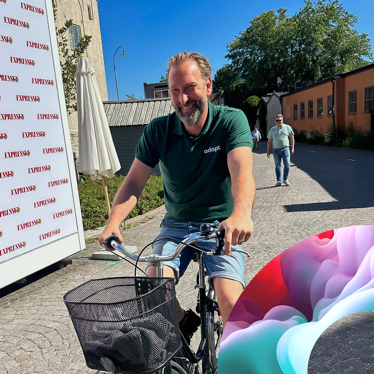 A smiling man rides a bicycle on a sunny cobblestone street, wearing a green polo and shorts. Another person walks in the background. Bright buildings and greenery are visible, with a colorful abstract shape in the lower right corner.