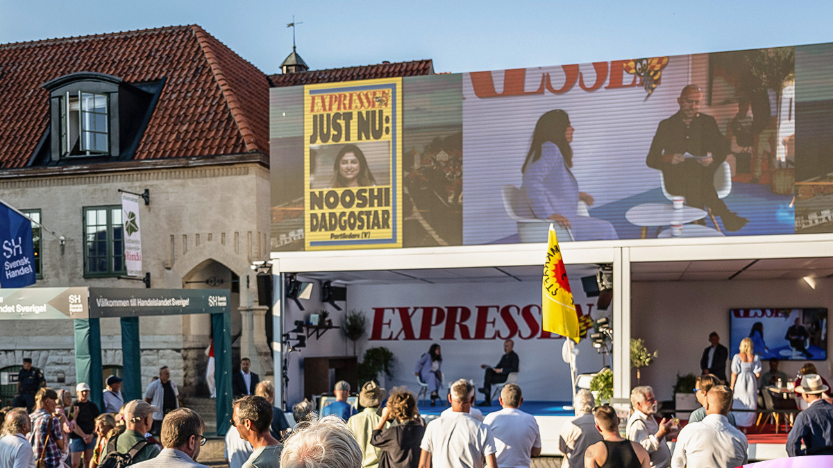 A crowd gathers outside a building with an EXPRESSEN banner, watching a large screen showing an interview with Nooshi Dadgostar. Colorful flags and banners are visible in the sunny outdoor setting.