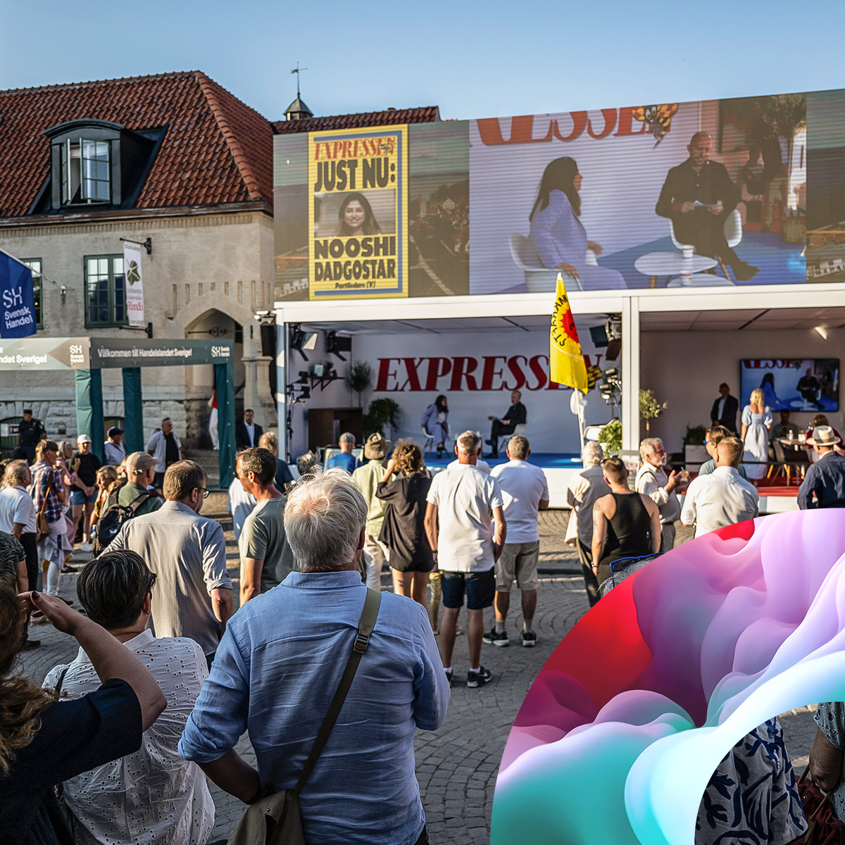 A crowd gathers outdoors in front of a stage with a large screen displaying a talk show. Colorful abstract shapes appear in the foreground. Buildings with red roofs are seen in the background.