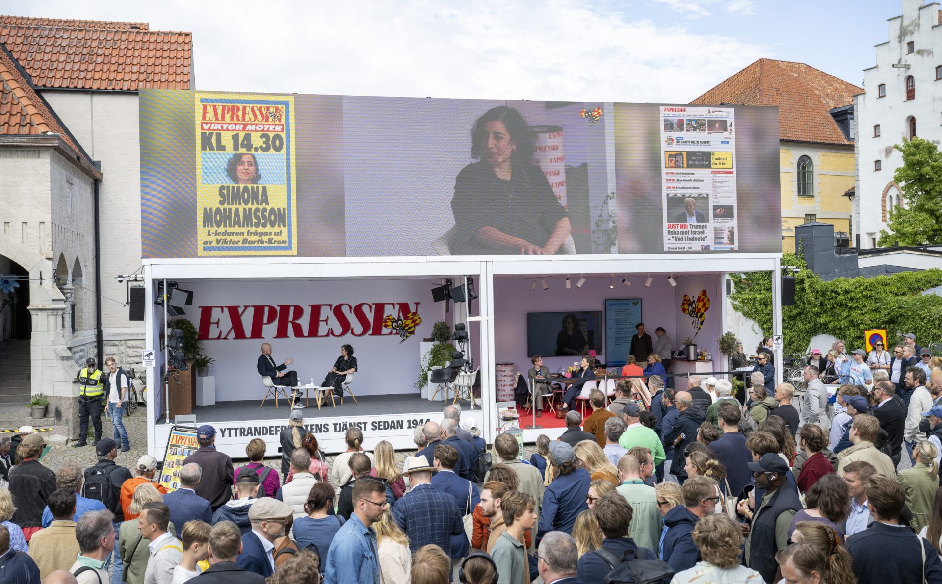 A large crowd gathers around an outdoor stage with “EXPRESSEN” banners, where a panel discussion is taking place. A large screen above displays a close-up of a woman speaking, with news graphics and event details.