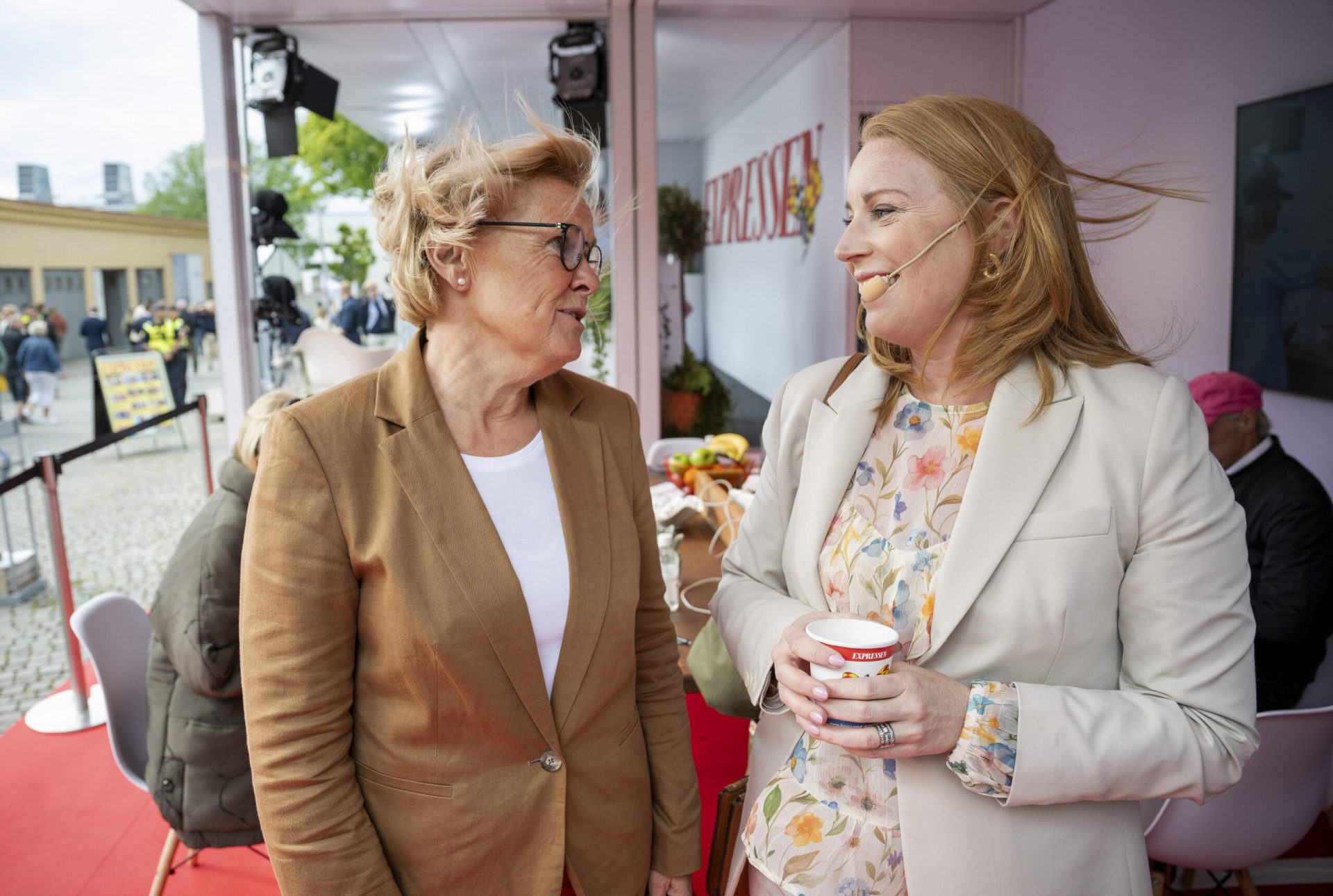 Two women stand and talk inside a tented area. Both are smiling; one wears a brown blazer and glasses, the other a cream blazer with a floral shirt, holding a cup and wearing a headset microphone. People are seated in the background.