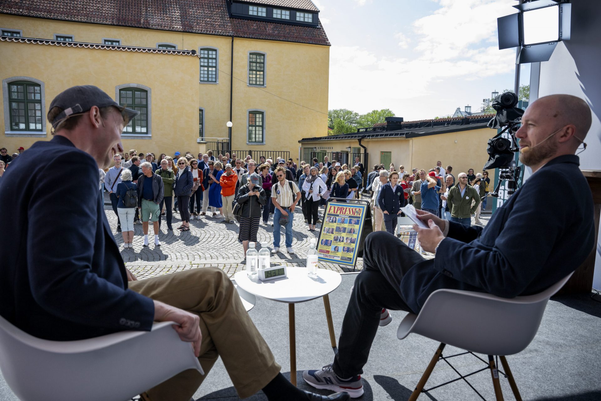 Two men sit on a stage in front of a crowd outdoors, engaged in a discussion. One holds a paper and smiles. A camera records the event, and a magazine is displayed between them. Bright, sunny day; historic buildings in background.