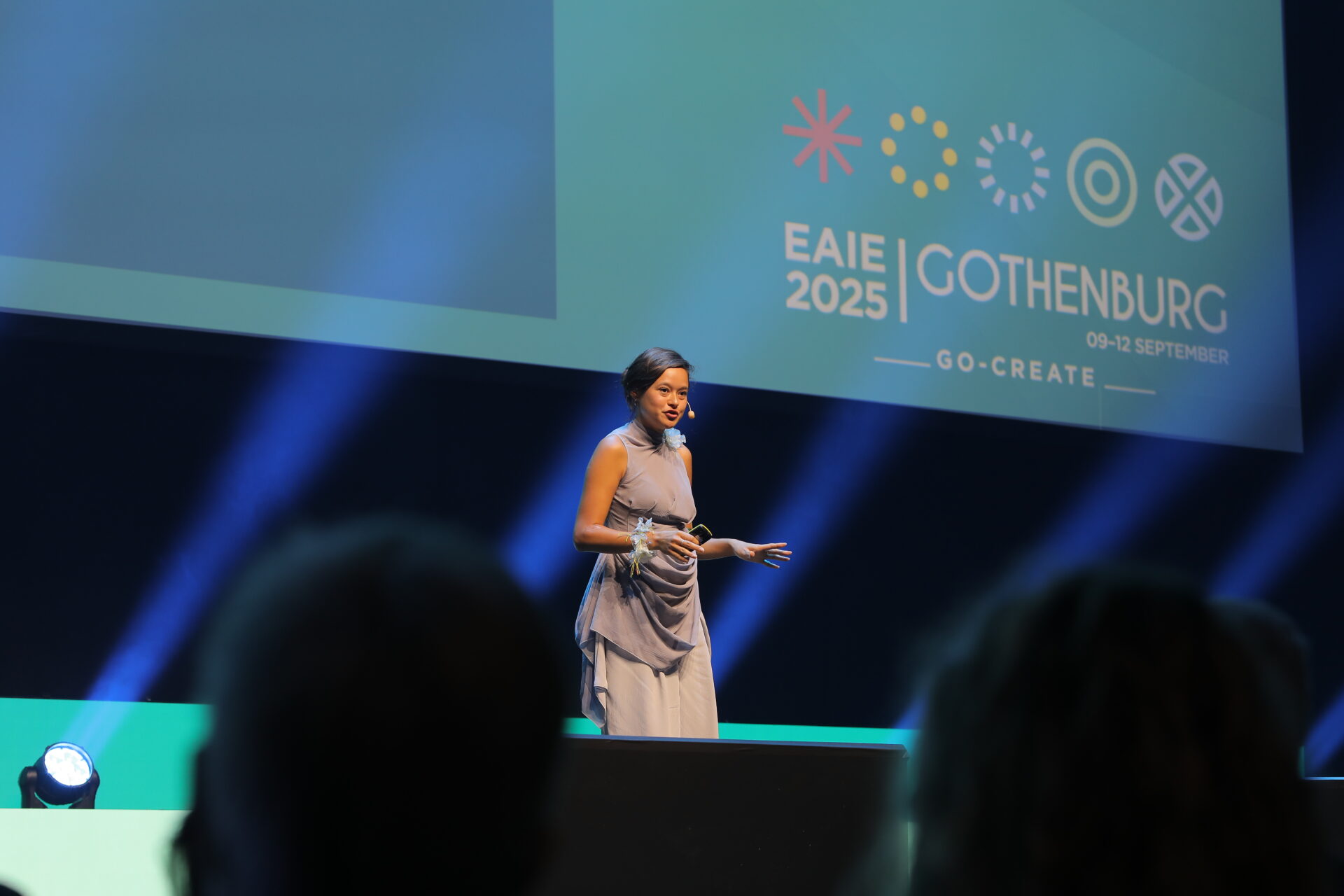 A woman stands on stage speaking at a conference, with EAIE 2025 Gothenburg 09-12 September and colorful symbols displayed on a large screen behind her. Audience members are visible in the foreground.