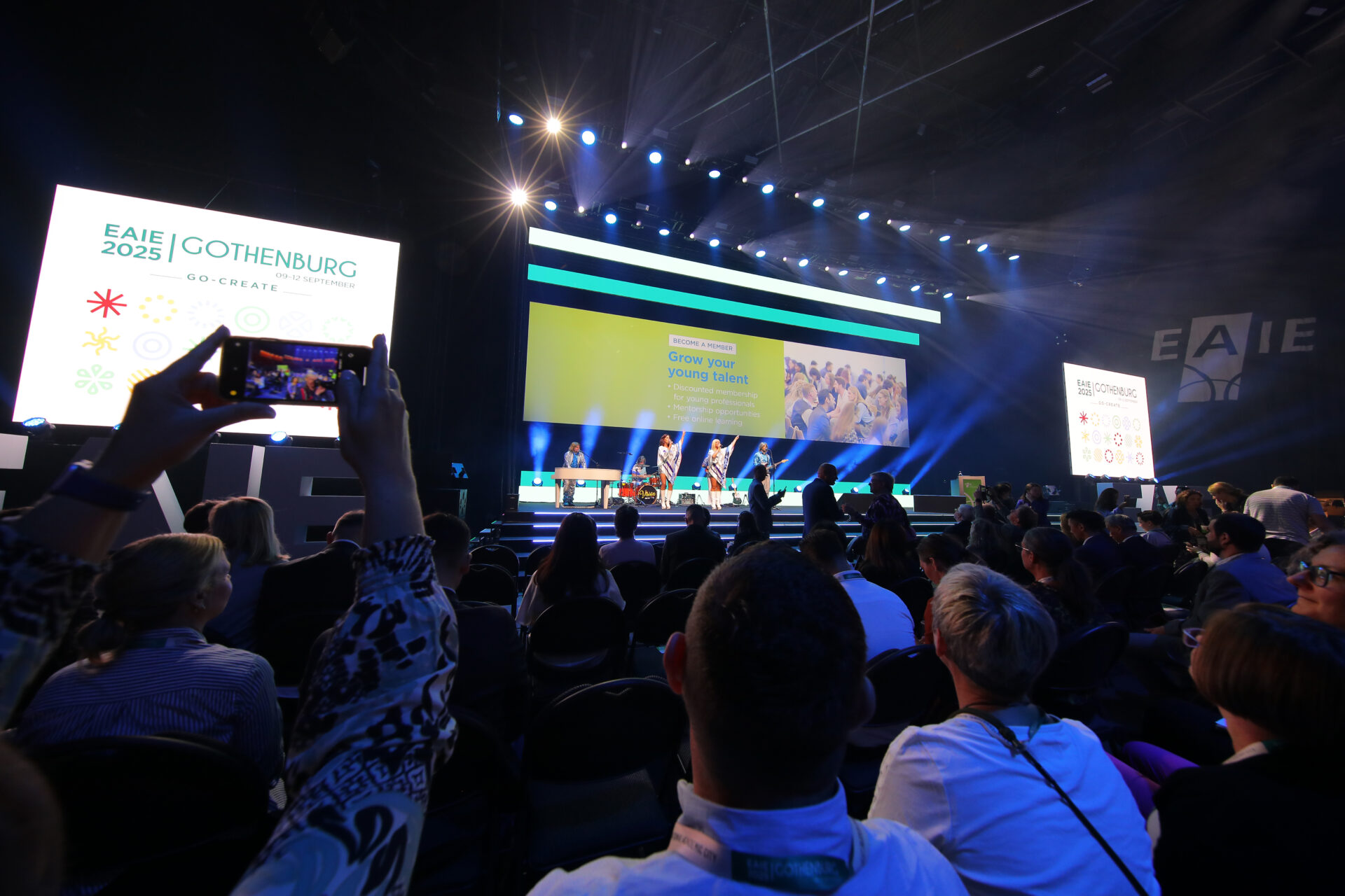 People seated in a large conference hall watch a brightly lit stage with panelists, large screens displaying EAIE Gothenburg 2025 and Grow your global future, and audience members taking photos.