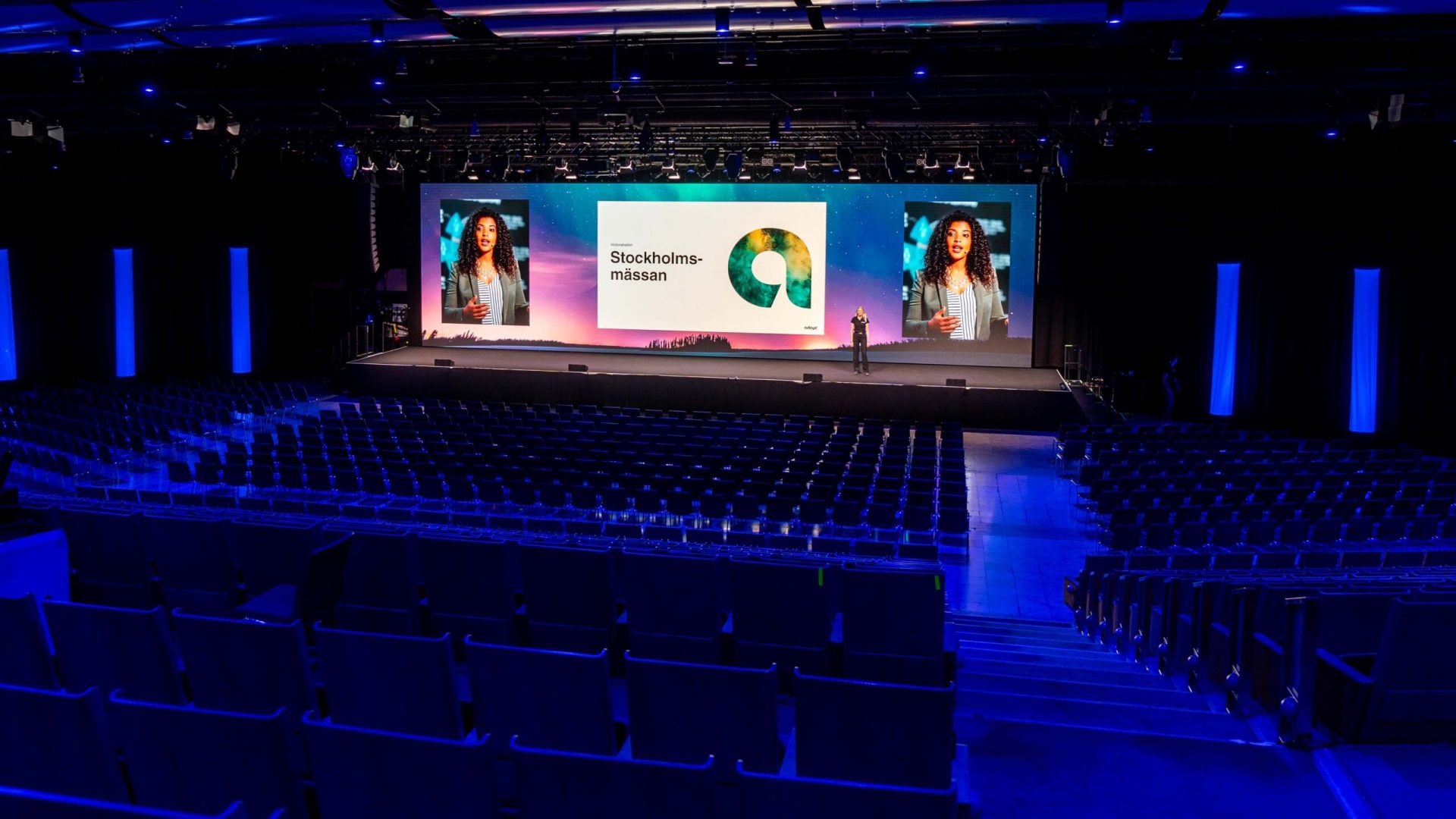 A large, modern auditorium with rows of empty seats facing a brightly lit stage. A presenter stands on stage near a screen displaying Stockholmsmässan and colorful graphics, with two large portraits on either side.