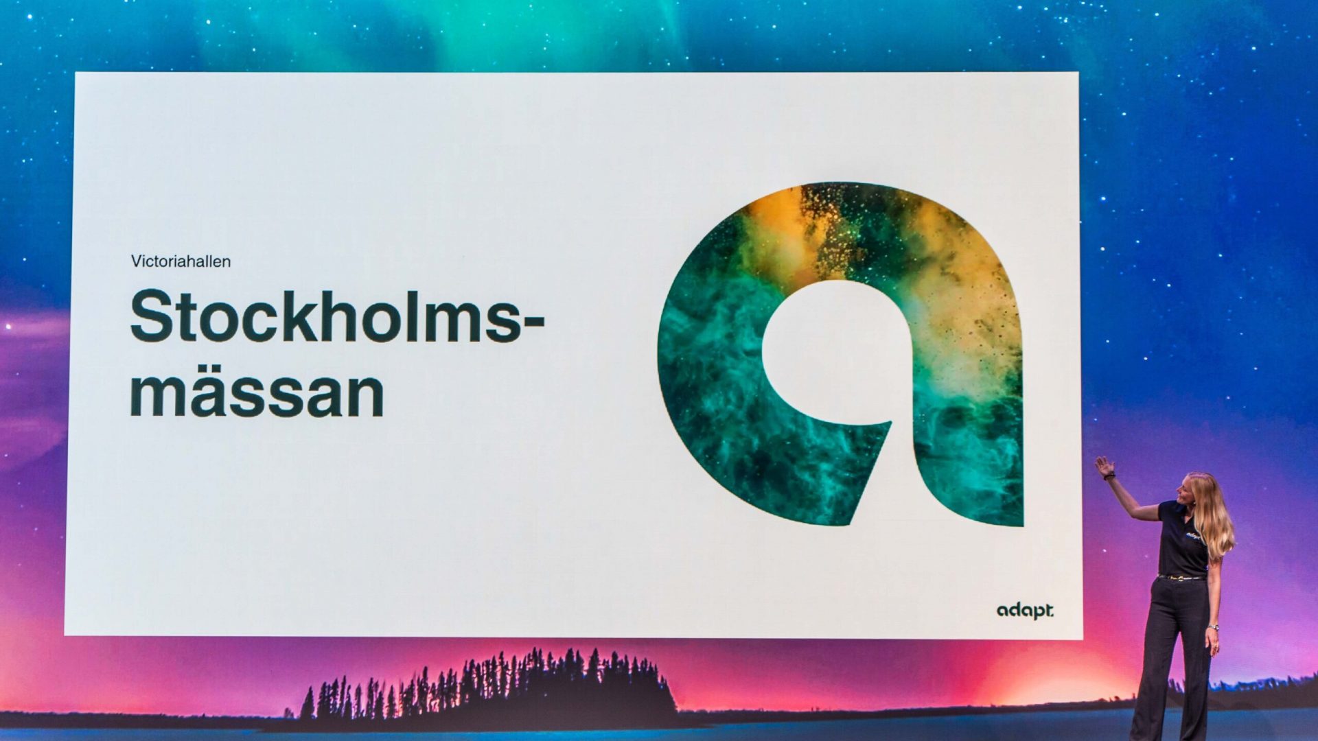 A woman stands on stage pointing to a large screen displaying the text Victoriahallen Stockholmsmässan and a stylized letter a with a colorful pattern, against a backdrop of a night sky and tree silhouettes.