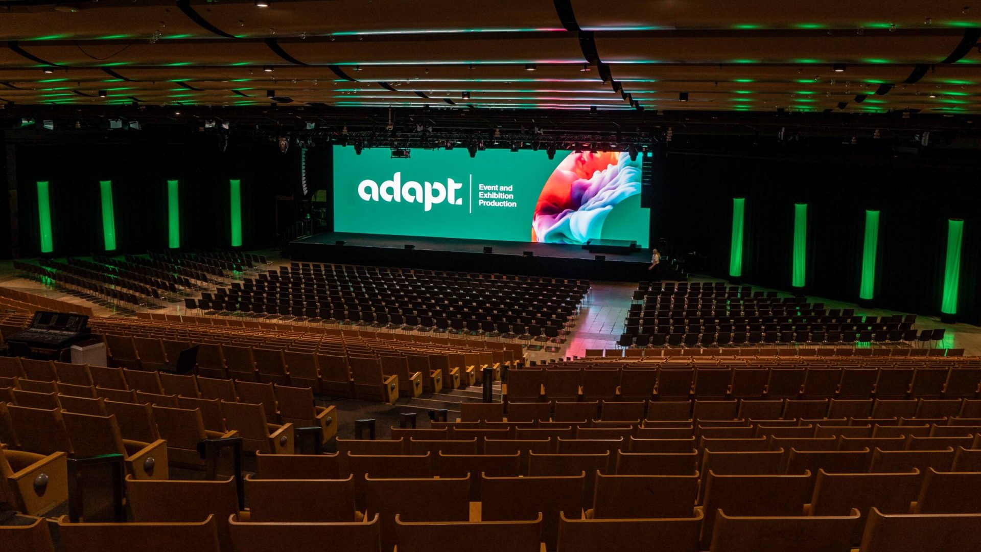 A large, empty auditorium with rows of brown seats facing a stage. The stage has a large green screen displaying adapt Event and Exhibition Production with colorful graphics. Green lighting accents the walls and ceiling.